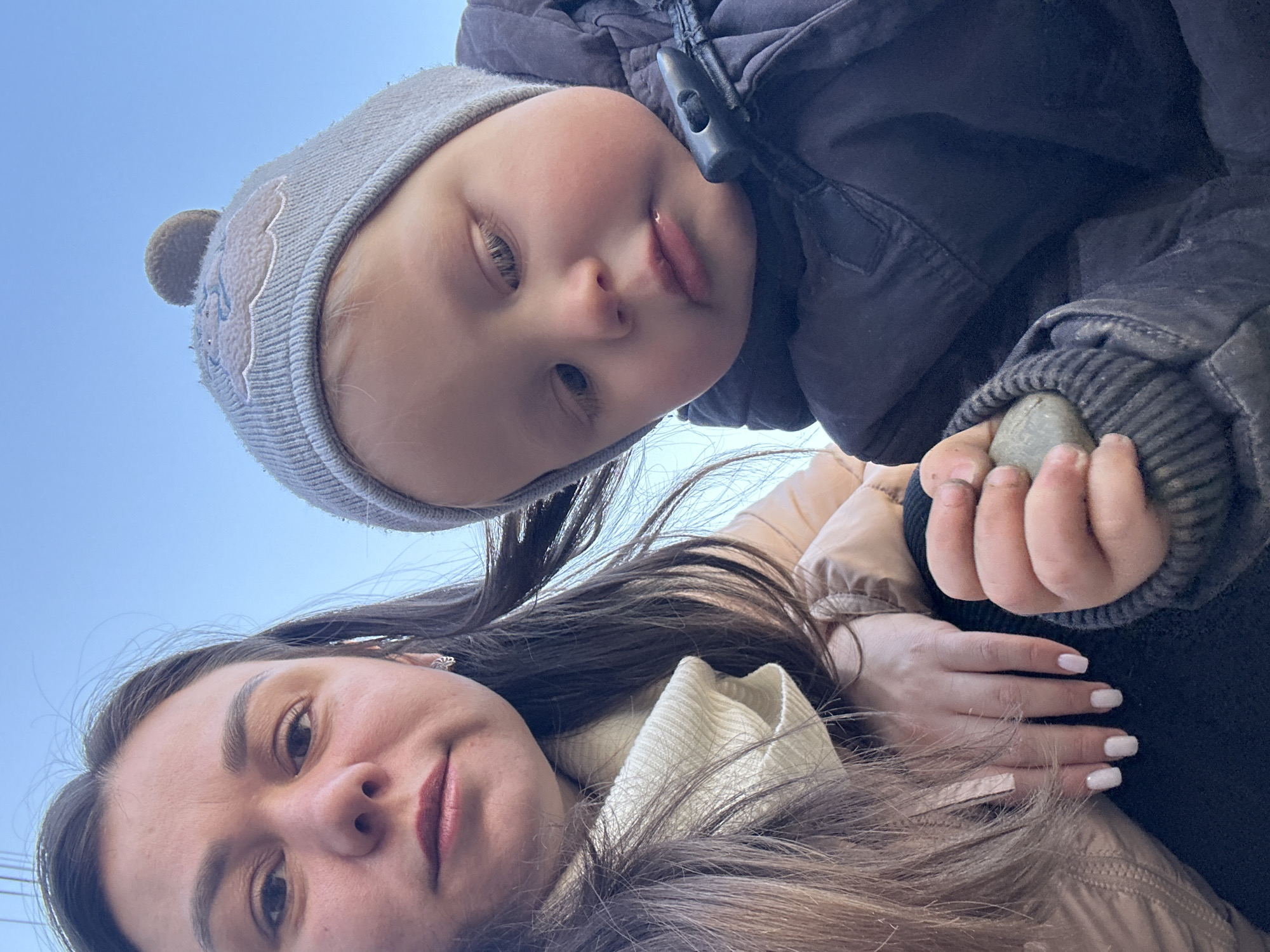 A low-angle, upward-facing selfie features a woman and a young child outdoors in Chaikovsky, Russia. The woman, with light skin, brown hair, and light-colored lipstick, gazes at the camera. The child, positioned above her and slightly to the right, wears a grey knit hat with a pom-pom and a dark grey winter coat. The child's right hand, visible between the child and the woman, holds a small grey rock. The sky, a light blue, is visible in the background. The woman wears a cream-colored turtleneck sweater and a tan-colored jacket. The child's coat has a toggle closure. The woman's nails are painted a light color.