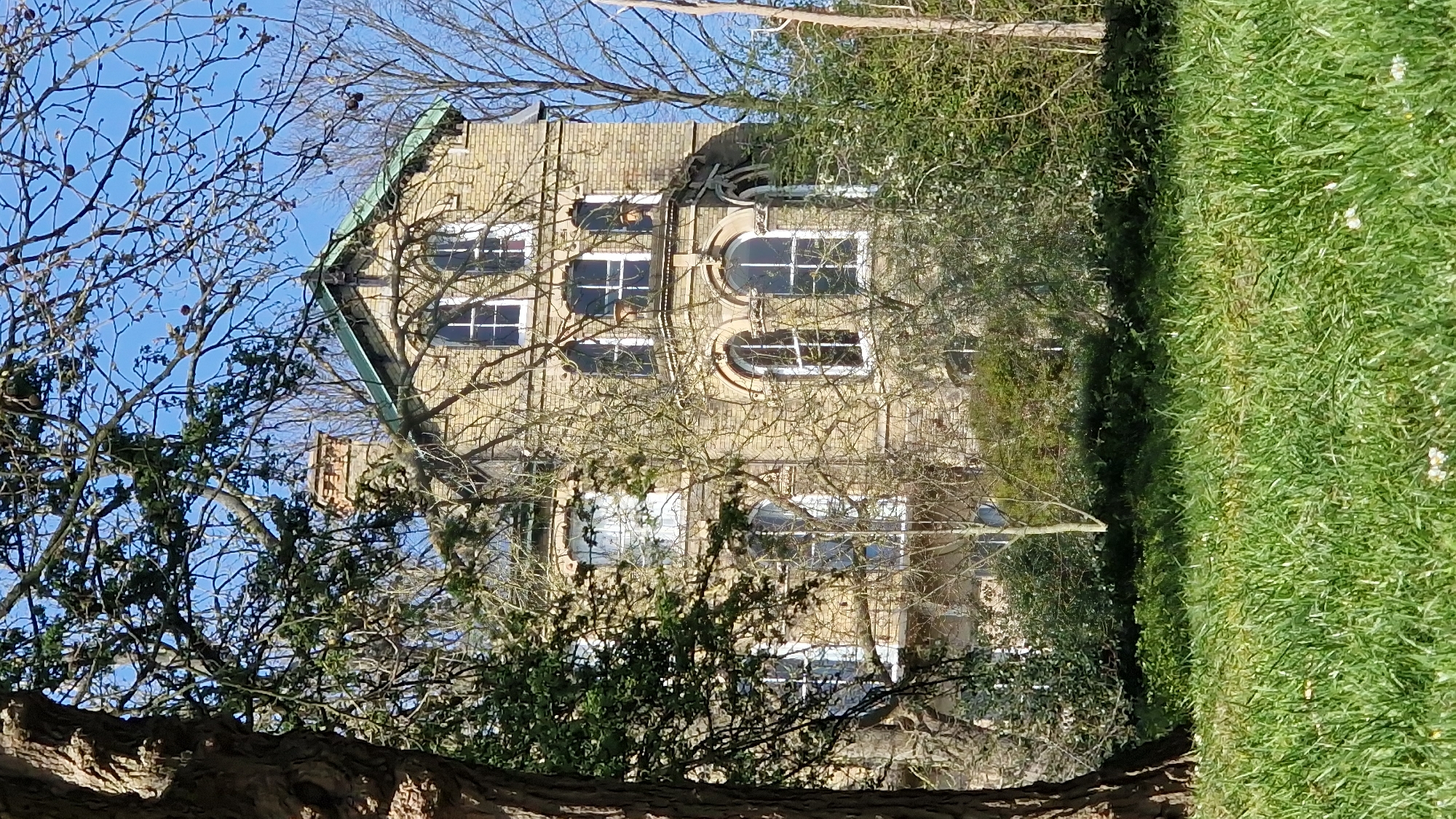 A three-story brick building is partially obscured by trees. The building is light brown in color and has a green roof. It features rectangular and arched windows. The surrounding environment includes green grass and trees with bare branches. The image was likely taken in the city of Lincoln, United Kingdom.
