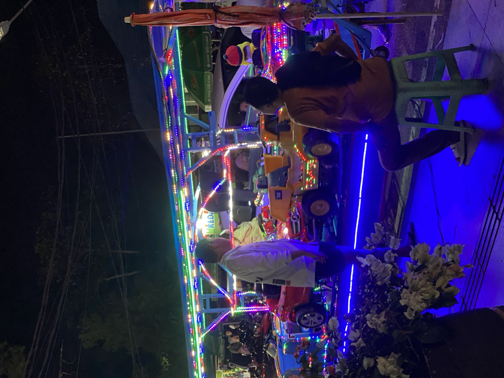 The image depicts an outdoor night scene in the city of Banjarbaru, Indonesia, likely a recreational area with amusement rides. In the foreground, a person sits on a green plastic stool facing away from the camera, wearing an orange t-shirt and dark pants. To the left, a person in a white long-sleeved shirt and dark shorts stands looking towards a row of brightly lit toy cars. These cars, a red Mercedes-Benz and a yellow construction vehicle, are illuminated with strings of colorful LED lights, including green, blue, and purple. In the background, a white car is parked, and other people are visible in the distance. Overhead, a structure adorned with more colorful lights supports a blue awning. Green umbrellas with white lettering are also visible. The ground is paved with dark tiles, and the overall atmosphere is festive due to the extensive use of decorative lighting.
