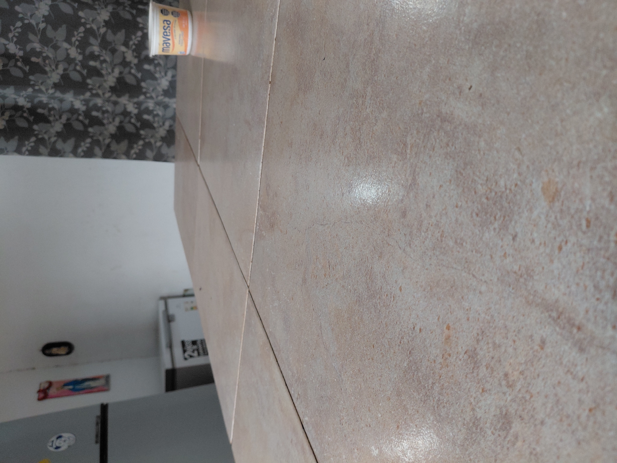 The image, oriented vertically, depicts an indoor scene, likely in a kitchen or utility area in Santa Ana, Venezuela. The foreground and right side of the frame are dominated by a glossy, light brown or beige tiled surface, which appears to be a countertop or floor. These tiles feature subtle variations in tone and small, darker speckles, resembling natural stone, and are divided by visible grout lines.
Lying horizontally on this tiled surface in the midground is a small, cylindrical plastic container. Its label clearly shows the brand name "mavesa" in distinct orange lettering on a white background, suggesting it is a food product such as margarine or a dairy item.
The background, positioned behind the tiled surface, reveals various interior elements. To the far left, a dark curtain or fabric with a light-colored, leafy floral pattern is visible. Below this, a plain white wall is seen, upon which a small, dark oval object is mounted. Further down on the wall, a framed picture or artwork depicting a religious figure, possibly a Madonna or saint, is displayed. Towards the bottom left, the top section of a grey or silver refrigerator with a sticker attached to it is partially visible. Beyond this, further into the background, another white appliance, likely a second refrigerator or tall cabinet, stands with some indistinct black text on its side.
The scene is well-lit, with reflections on the tiled surface indicating an indoor light source. There are no people present, and no specific activity is occurring.