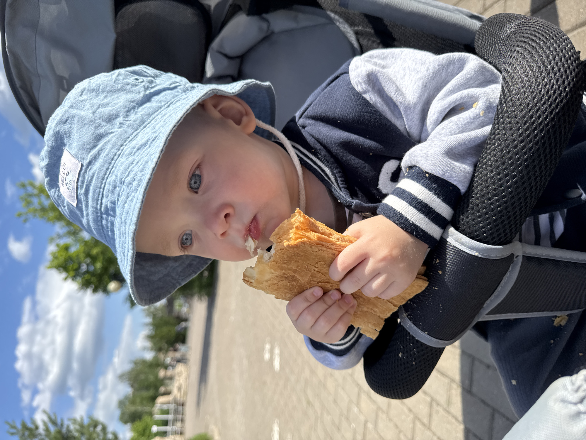 A toddler wearing a light blue bucket hat and a navy and grey jacket sits in a stroller and eats a piece of bread. The child has blue eyes and is looking to the right of the frame. The stroller has a black mesh armrest. In the background, a paved walkway and trees are visible under a partly cloudy blue sky. The image was taken in Izhevsk, Russia, at an outdoor location, possibly a park or a public garden.