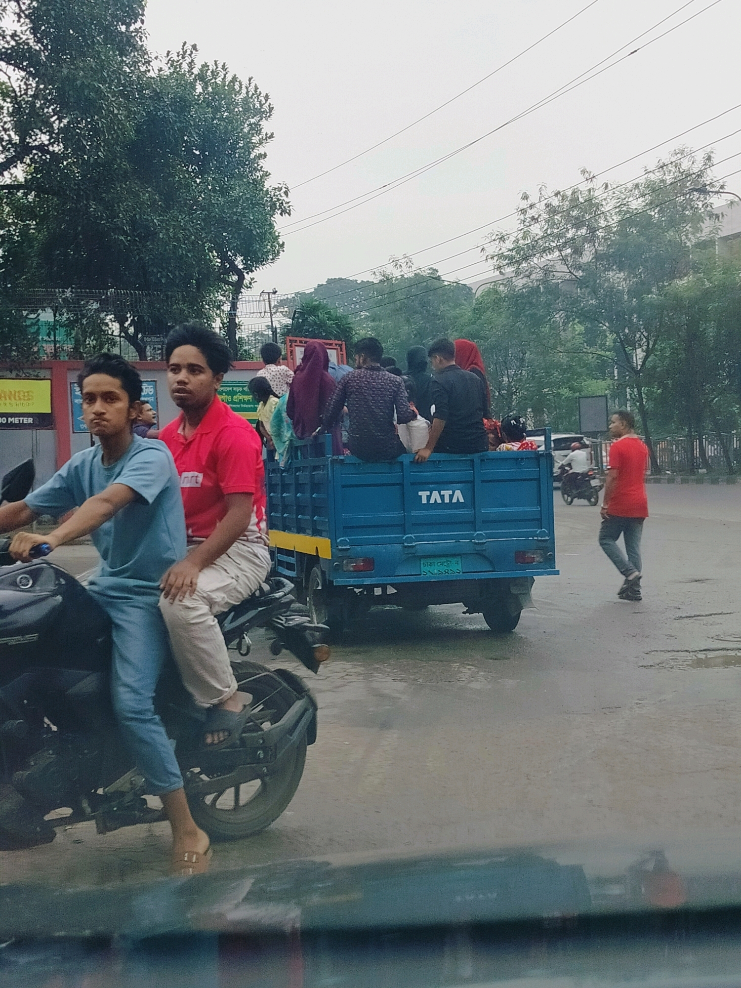 This image captures a scene on a city street in Dhaka, Bangladesh, likely during overcast or rainy weather as indicated by the wet road surface. In the foreground, two young men are riding a motorcycle. The rider, wearing a light blue t-shirt and pants, has his hands on the handlebars. The pillion rider, in a red polo shirt and light-colored pants, sits behind him, looking forward with a slightly anxious expression.
Behind the motorcycle, a blue Tata truck is visible, transporting several passengers in its open bed. Among them, a person in a red hijab is prominent, along with others dressed in various casual attire. The truck bears the "TATA" logo on its side.
Further down the street, another man in a red t-shirt and jeans is walking, looking towards the truck. A motorcycle is also seen in the background to the right, carrying a rider. Trees line the street, and the overall environment suggests a busy urban setting. The presence of utility wires overhead is also a notable environmental detail. The overall impression is one of everyday transit and crowded conditions in Dhaka.