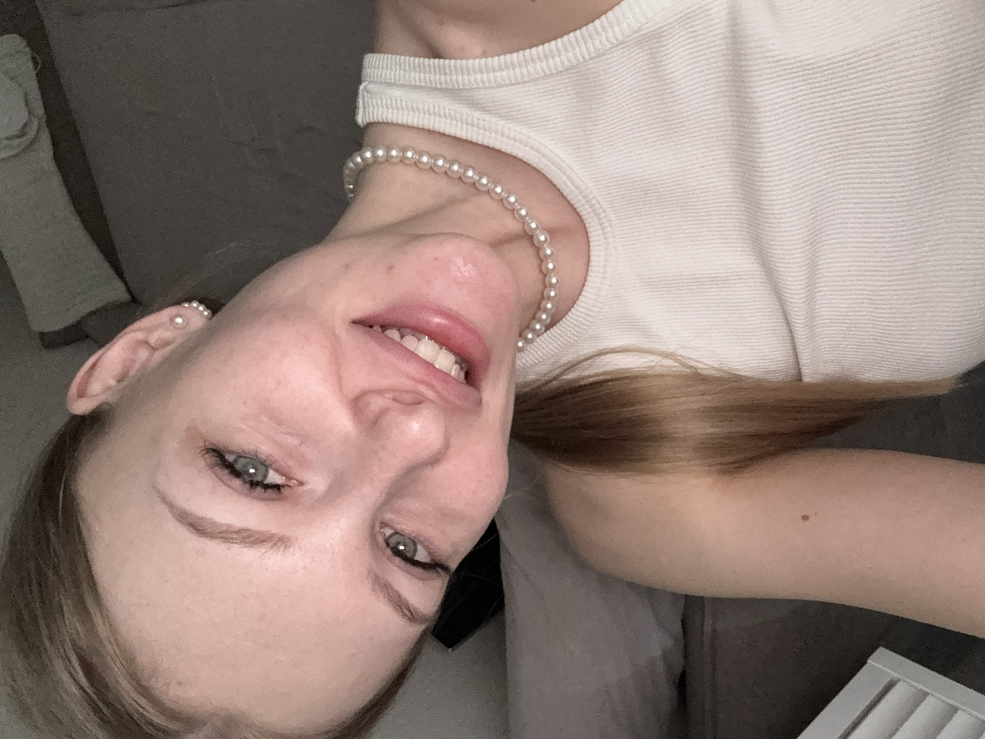 A woman with fair skin, light brown hair, and blue eyes is smiling at the camera. She is wearing a white ribbed tank top and a pearl necklace. There is a pearl earring in her left ear. The background appears to be a gray couch. The location is not discernable in the image.