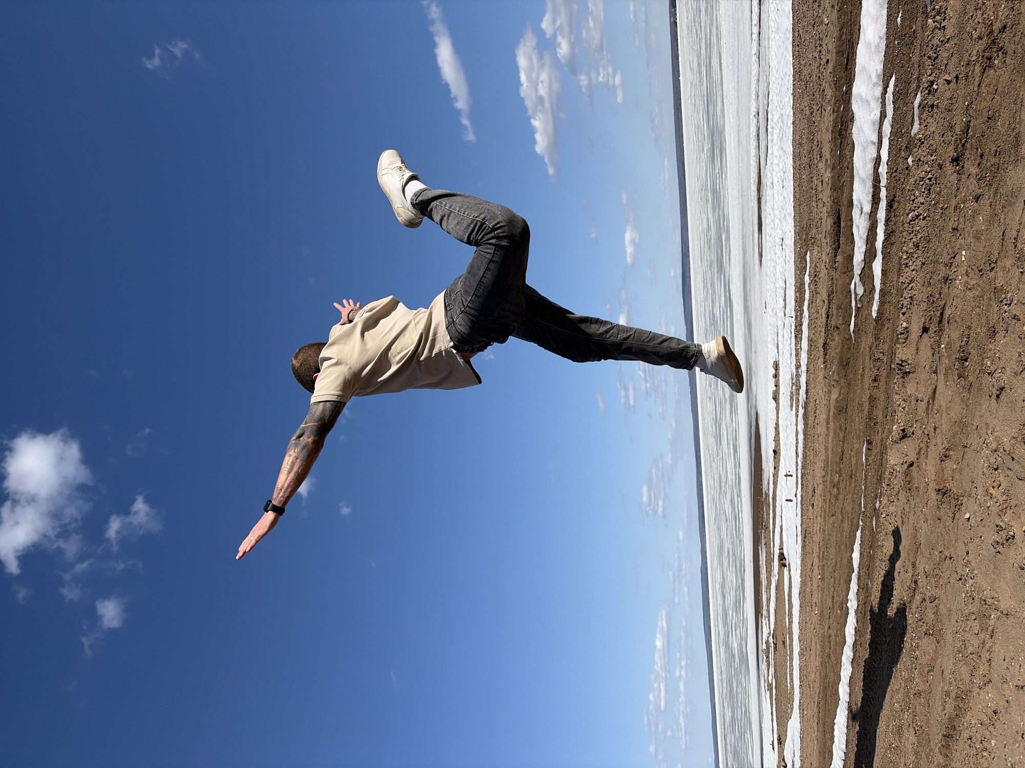 A man is suspended upside down against a backdrop of a bright blue sky with scattered white clouds. He wears a tan shirt, dark jeans, and light-colored shoes. Tattoos are visible on his extended arm. Below him is a horizontal landscape showing a partially frozen lake or body of water bordered by a sandy or dirt-covered bank with patches of snow. The image is taken with the horizon line running vertically on the right side of the frame. The location is Noviy, Russia.