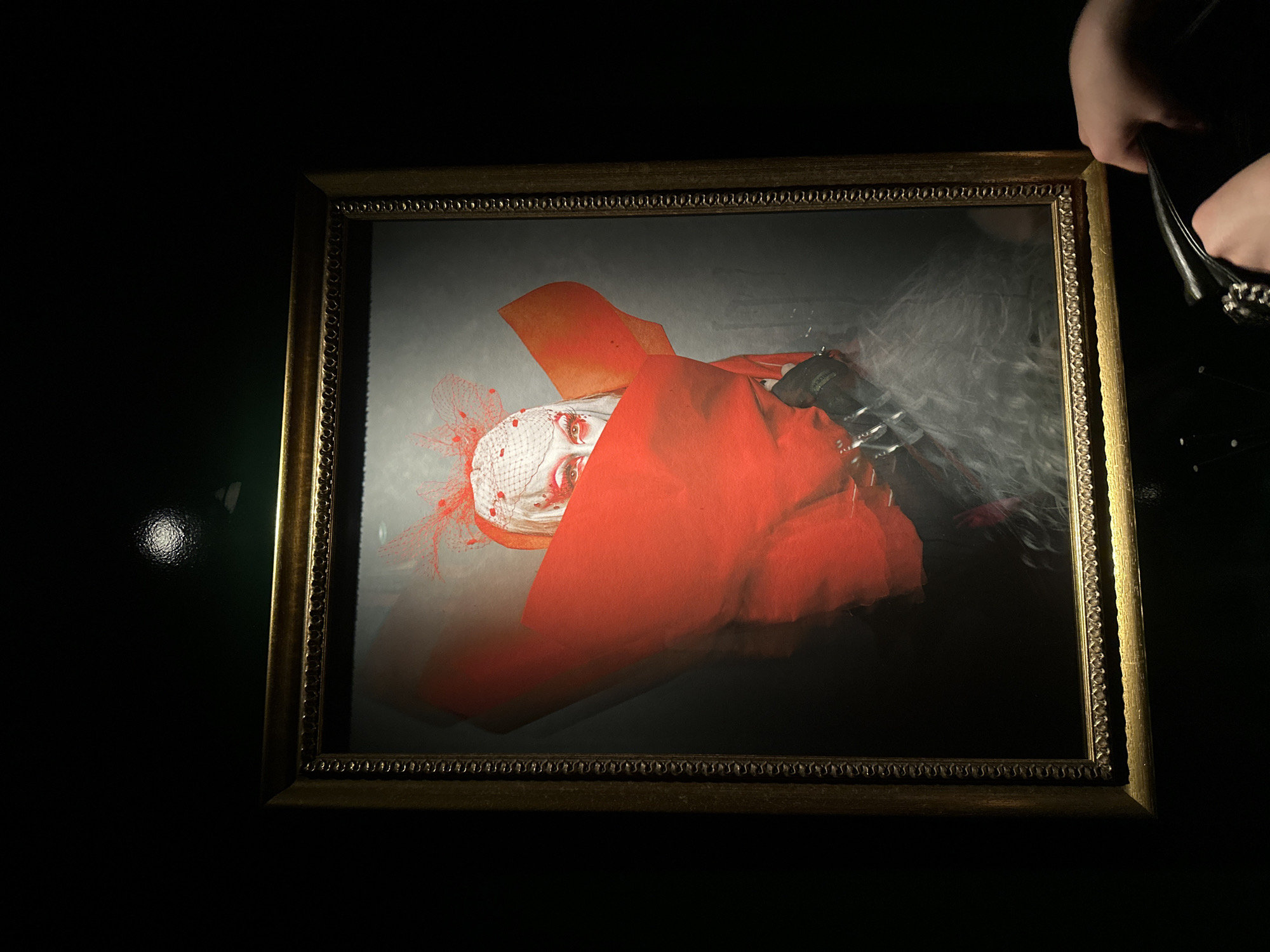 The photograph shows a framed image hanging on a dark wall. The frame is ornate, gold-colored, and rectangular. The image inside the frame appears to be a portrait of a person wearing a red garment and a white head covering with red netting. A pair of hands, possibly holding a black object or accessory, is visible in the upper right corner of the frame. The environment is dimly lit, creating a stark contrast between the brightly lit artwork and the surrounding darkness. The scene is not explicitly linked to London or the United Kingdom.