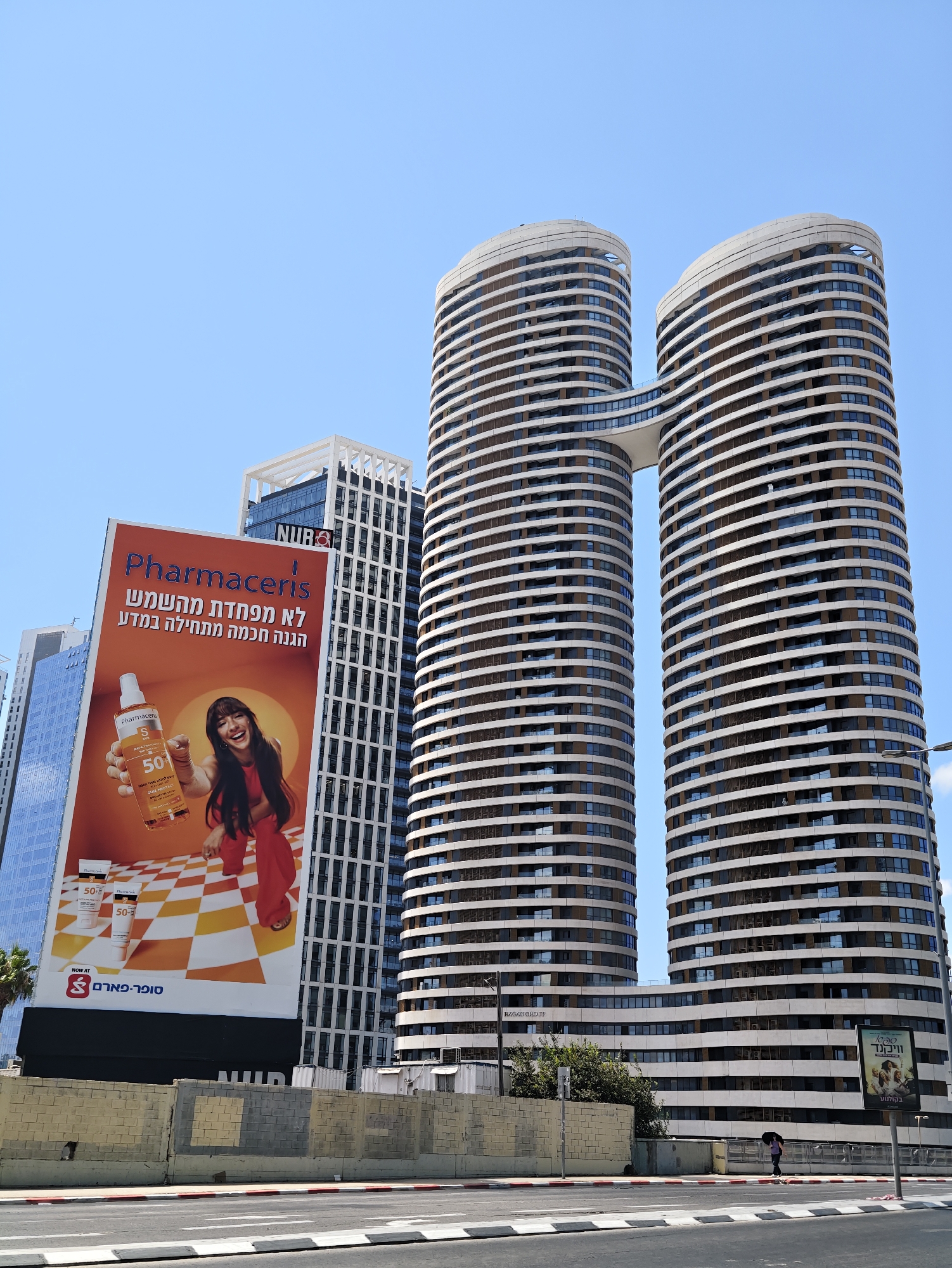 In Givatayim, Israel, two distinctive, curved skyscrapers, connected by a skybridge, dominate the skyline under a clear blue sky. The skyscrapers feature horizontal banding and numerous windows. In the foreground, a large billboard advertises "Pharmaceris" sunscreen with an image of a woman holding a product and text in Hebrew. To the left and behind the billboard, other modern buildings are visible, including one with a white, grid-like facade. A street with crosswalks runs in front of the buildings. A person is walking with an umbrella on a sidewalk to the right of the scene, and a street lamp is visible. A low, light-colored wall runs along the sidewalk in the foreground.