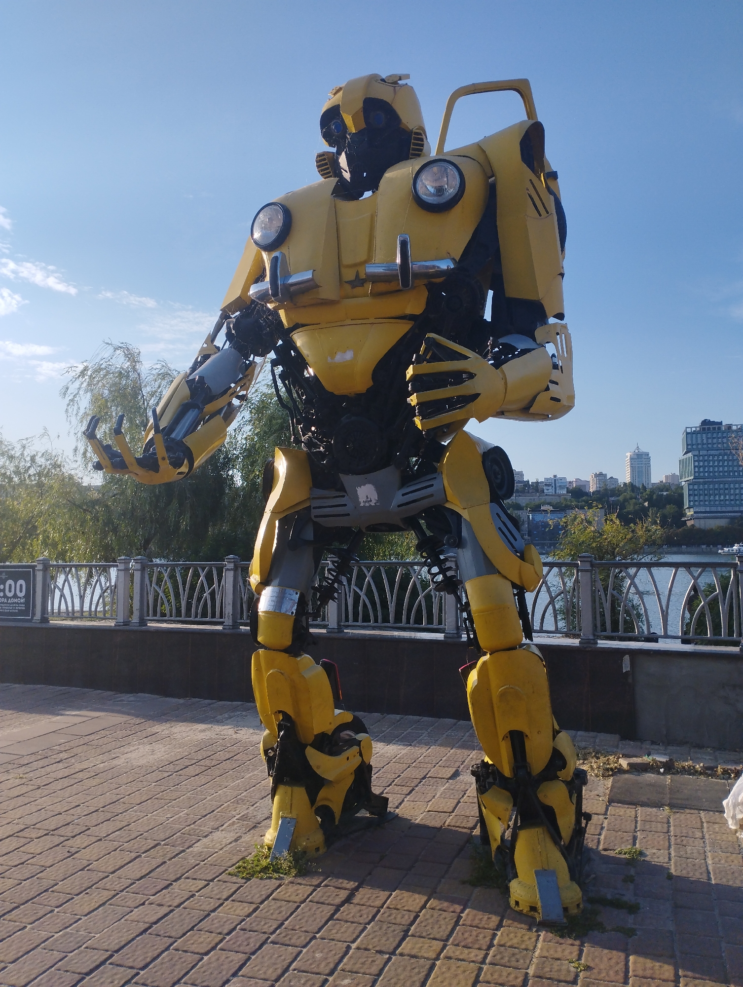 A large, yellow robot resembling the character Bumblebee from Transformers stands on a paved walkway next to a decorative metal fence. The robot is made of scrap metal and car parts, with visible mechanical joints and exposed wires. It is positioned in an outdoor setting with a clear blue sky and some clouds above. In the background, there are trees, a body of water, and a city skyline with several buildings. To the left of the robot, a sign with Cyrillic text and a clock displaying "0:00" is visible. The robot is the central subject of the image. The location is identified as the embankment in Rostov-on-Don, Russia.