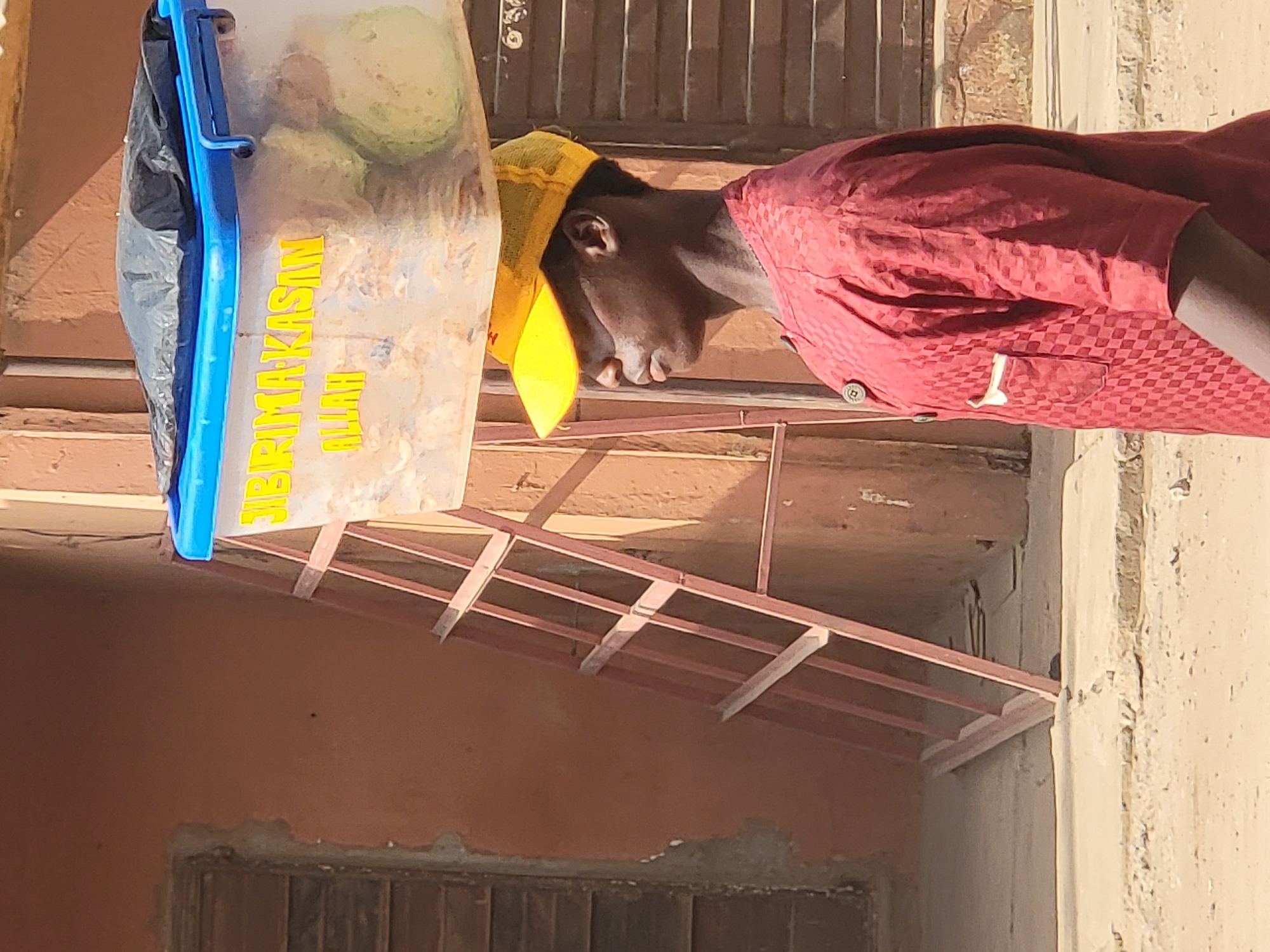 A man in a maroon checkered shirt and yellow head covering is carrying a blue-trimmed container. The container appears to hold food waste and has the words "ZIBIRIMA KASIM ALLAH" printed on it in yellow. The scene appears to be outside, possibly near a building with a rusty metal structure. The location is Maiburiji, Nigeria.