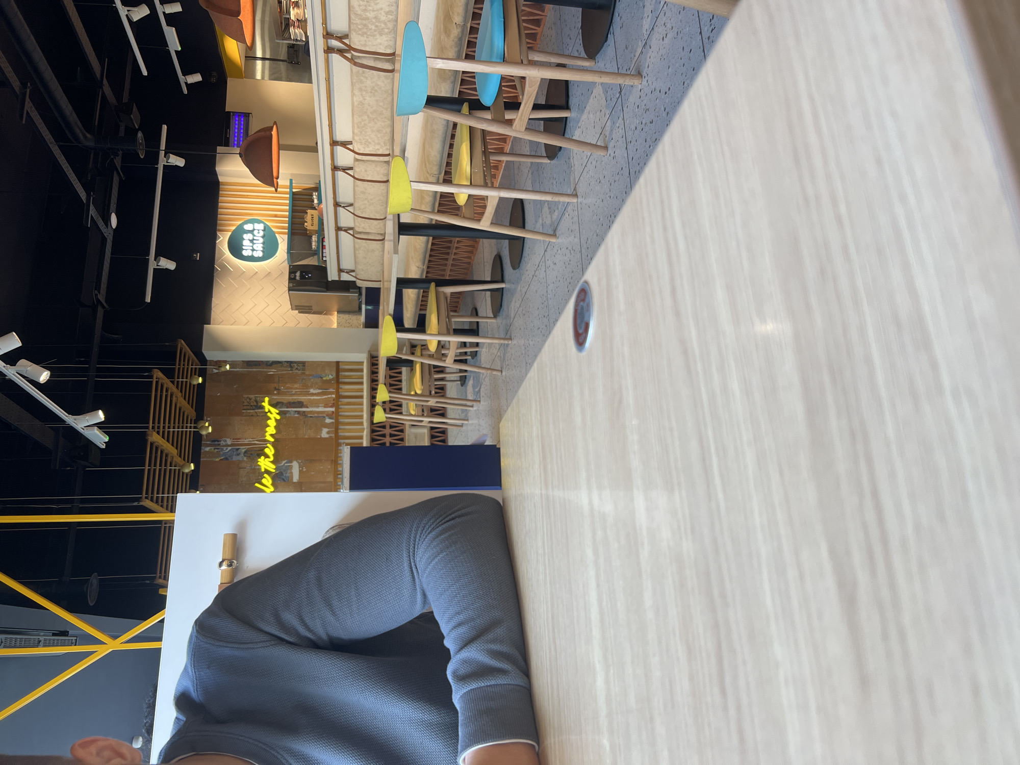 The image depicts the interior of "Sips & Sauce," a casual dining establishment located in Castle Quarter, Norwich, United Kingdom.
In the immediate foreground, a light-colored, subtly grained wooden table occupies the lower half of the frame. A circular metallic floor marker with a red center is partially visible on the tiled floor in the bottom left. On the left side, a person's torso and arm are visible, clad in a grey-blue textured long-sleeved sweater with a lighter cuff. The individual is seated at the table, with only the top of their right ear visible.
Beyond the foreground table, the restaurant features rows of light wooden tables with dark bases, accompanied by distinctive wooden chairs with bright yellow and teal/light blue seats and backs. Upholstered booth seating, covered in a light beige fabric with brown strap accents, runs along the back wall. The flooring throughout the visible area is a light grey speckled terrazzo or tiled surface. Overhead, a large, inverted rust-colored pendant lamp hangs, providing ambient light over one of the tables.
The background reveals the main service area of "Sips & Sauce." A prominent circular sign with a teal border and white background displays the restaurant's name. Below it, a counter holds a coffee machine or similar beverage dispenser and a small wooden box labeled "CLICK & COLLECT." The wall behind the counter is textured with a white brick pattern. To the far left, a feature wall made of vertical wooden slats includes a partial neon sign that reads "...le the most." The ceiling is industrial-style, painted black, with exposed pipes, ventilation ducts, and various track lighting fixtures. Decorative wooden slat structures also hang from the ceiling. The overall ambiance is bright and modern, with natural light entering from an unseen source on the right, creating a distinct shadow on the foreground table.