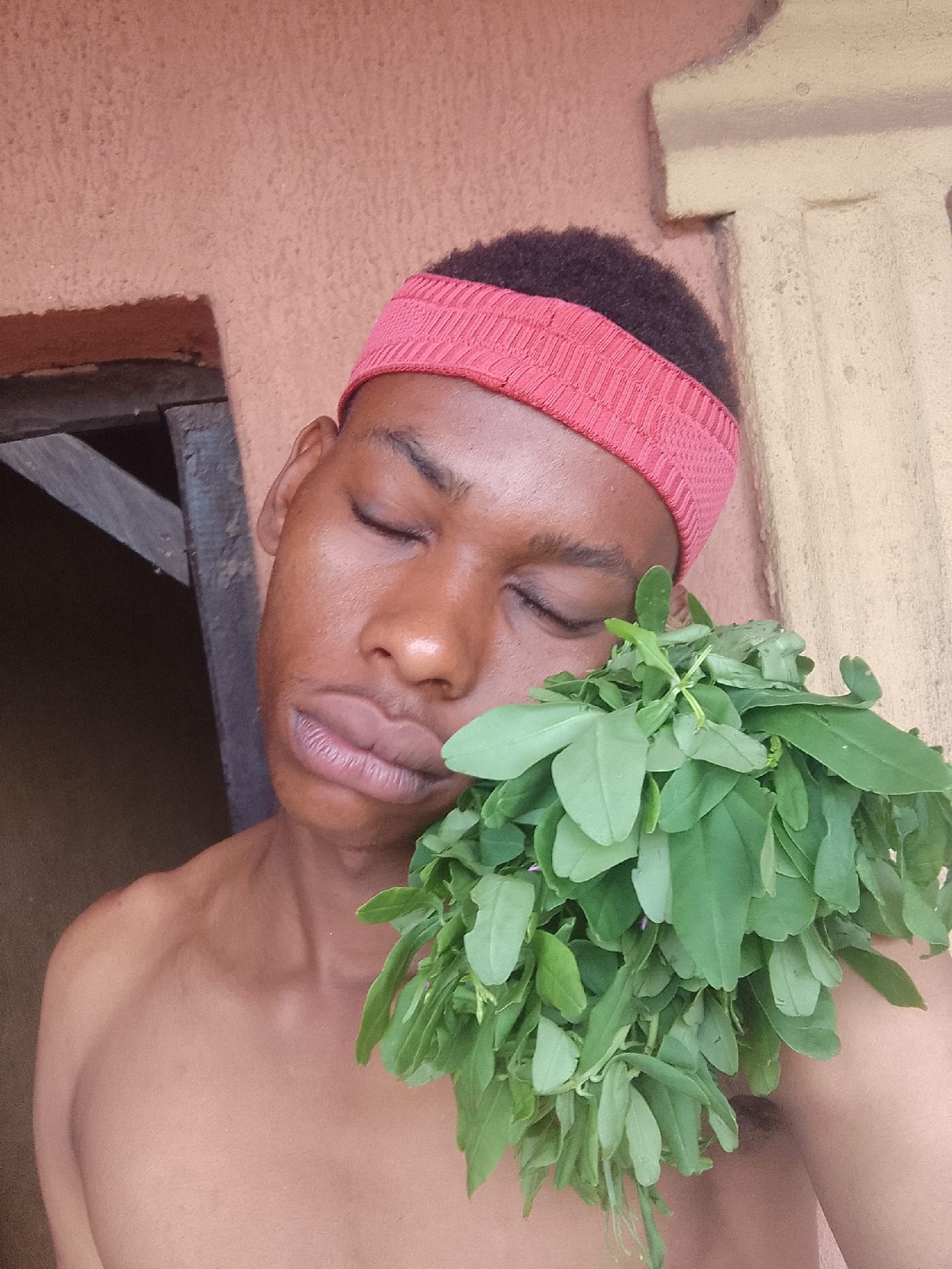 A shirtless man with dark skin and a red headband has his eyes closed and is holding a bunch of green leaves to his chest. The background shows a beige wall and part of a dark-colored doorway. Location: Unknown, Unknown.