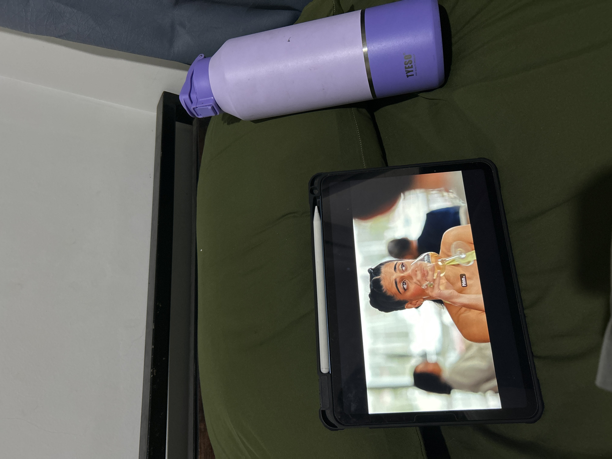 An overhead shot shows a tablet computer displaying a scene from a show with a woman drinking from a glass and a large purple and light purple water bottle on a dark green comforter. The tablet has a black case and a white stylus attached. The water bottle is a TYESO brand. The comforter is on a bed with a dark headboard visible. The background is a plain white wall. The location is not identifiable as Sukawati, Indonesia.