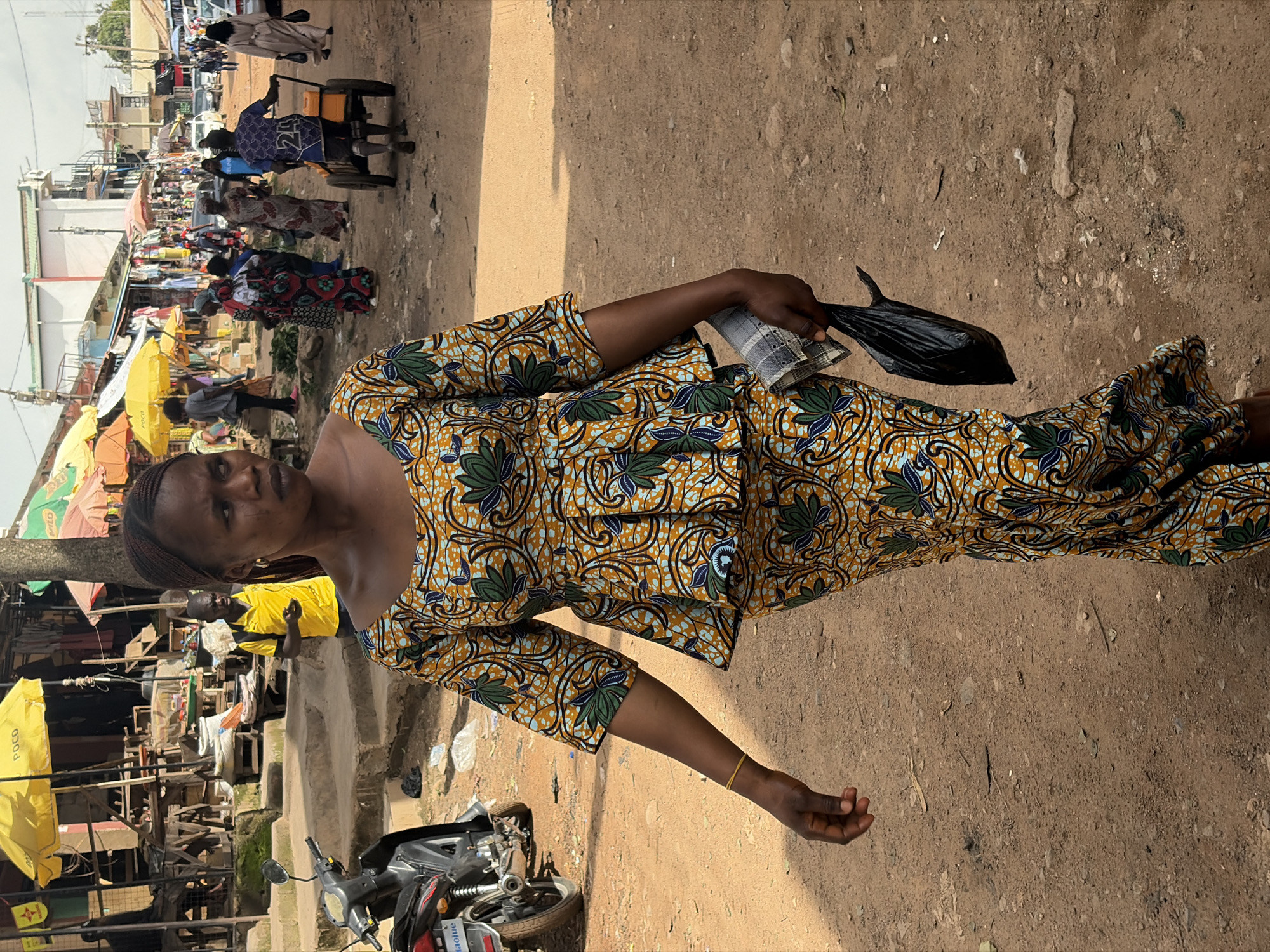 A woman is captured in a medium shot, standing in an outdoor market setting in Jos, Nigeria. She is wearing a vibrantly patterned yellow, blue, and green dress. Her left hand holds a black plastic bag, and her right arm is extended, with her hand open. The background depicts a bustling market scene with stalls, umbrellas, and various people, suggesting a lively commercial environment. A motorcycle is visible in the foreground to the left. The ground appears to be dirt or gravel, typical of an outdoor marketplace. The lighting suggests it is daytime, with sunlight casting shadows. The woman's expression is neutral. No visible text is discernable in the image.
