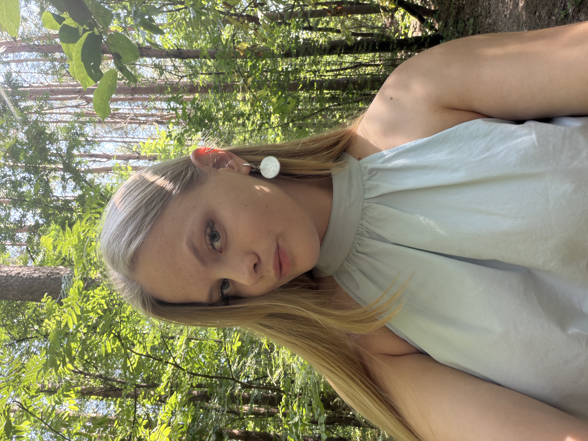 This media file is a portrait of a young Caucasian woman in an outdoor setting. The woman has blonde hair, blue eyes, and light skin. She is wearing a light blue top with a halter neckline. She is also wearing a circular white earring in her left ear. Her hair is styled in loose waves and falls around her shoulders.
The background is a forest with tall trees and dappled sunlight. The leaves on the trees are green and appear to be in full foliage, suggesting it is either spring or summer. The lighting indicates it is daytime, likely late morning or early afternoon, with the sun casting rays through the trees. The overall mood is serene and natural.
The woman is looking directly at the camera, with a relaxed and slightly contemplative expression. Her pose is informal, with her head tilted. The image appears to be a selfie, given the angle and framing.
The location context is Izhevsk, Russia. While there are no explicit text elements in the image, the visual cues of the foliage and tree types could potentially hint at a specific region within Russia. The natural environment and the woman's attire suggest a casual outing or a moment of relaxation in nature.