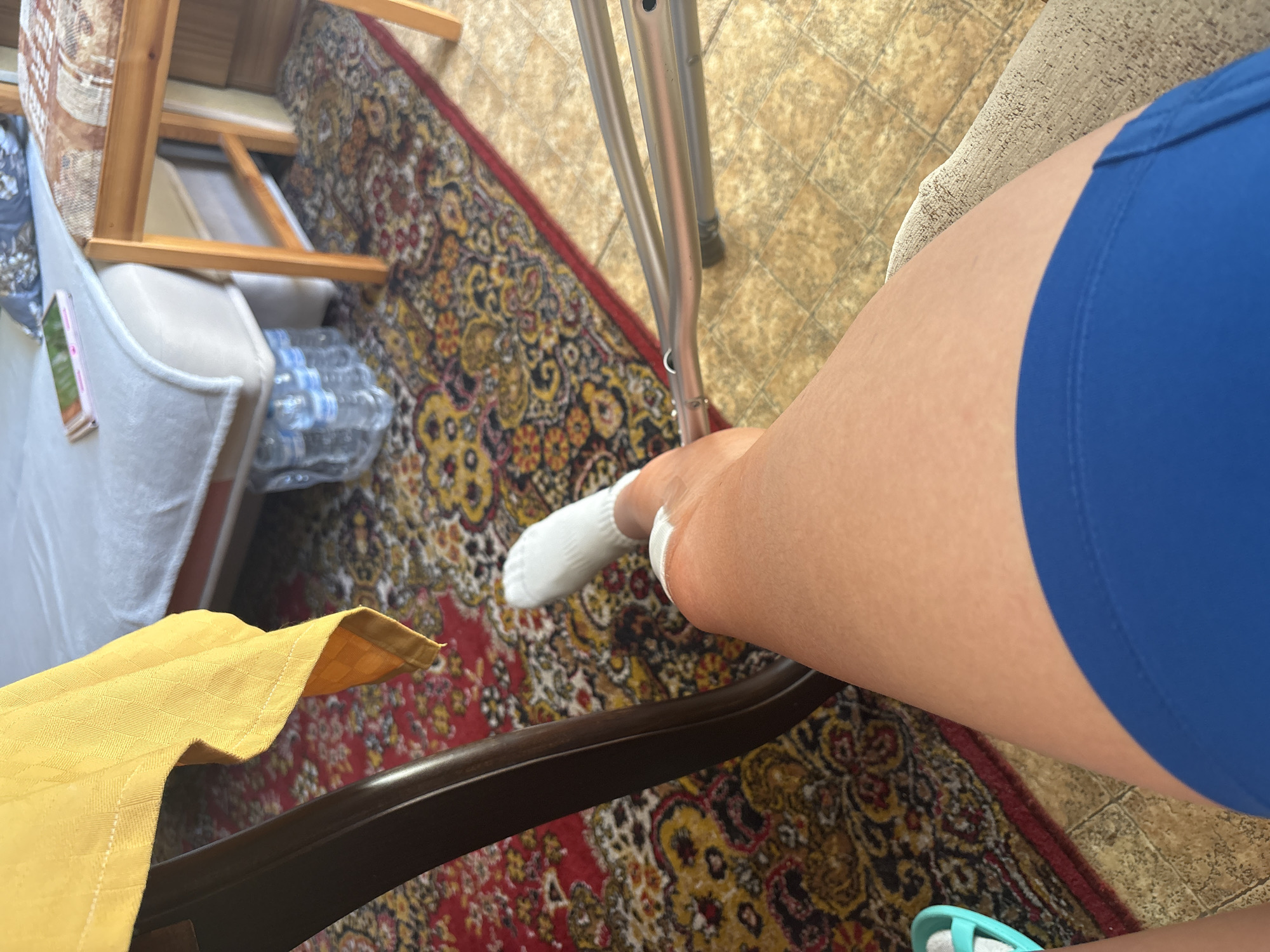 A first-person perspective image depicts a leg with a white sock and medical gauze around the ankle. The person is wearing blue shorts. A pair of crutches is partially visible in the background. The person is seated, with part of a chair visible in the foreground. The floor is covered with a patterned rug. A stack of bottled water is visible near a couch. The location is indoors, possibly in Sofia, Bulgaria, given the prompt.