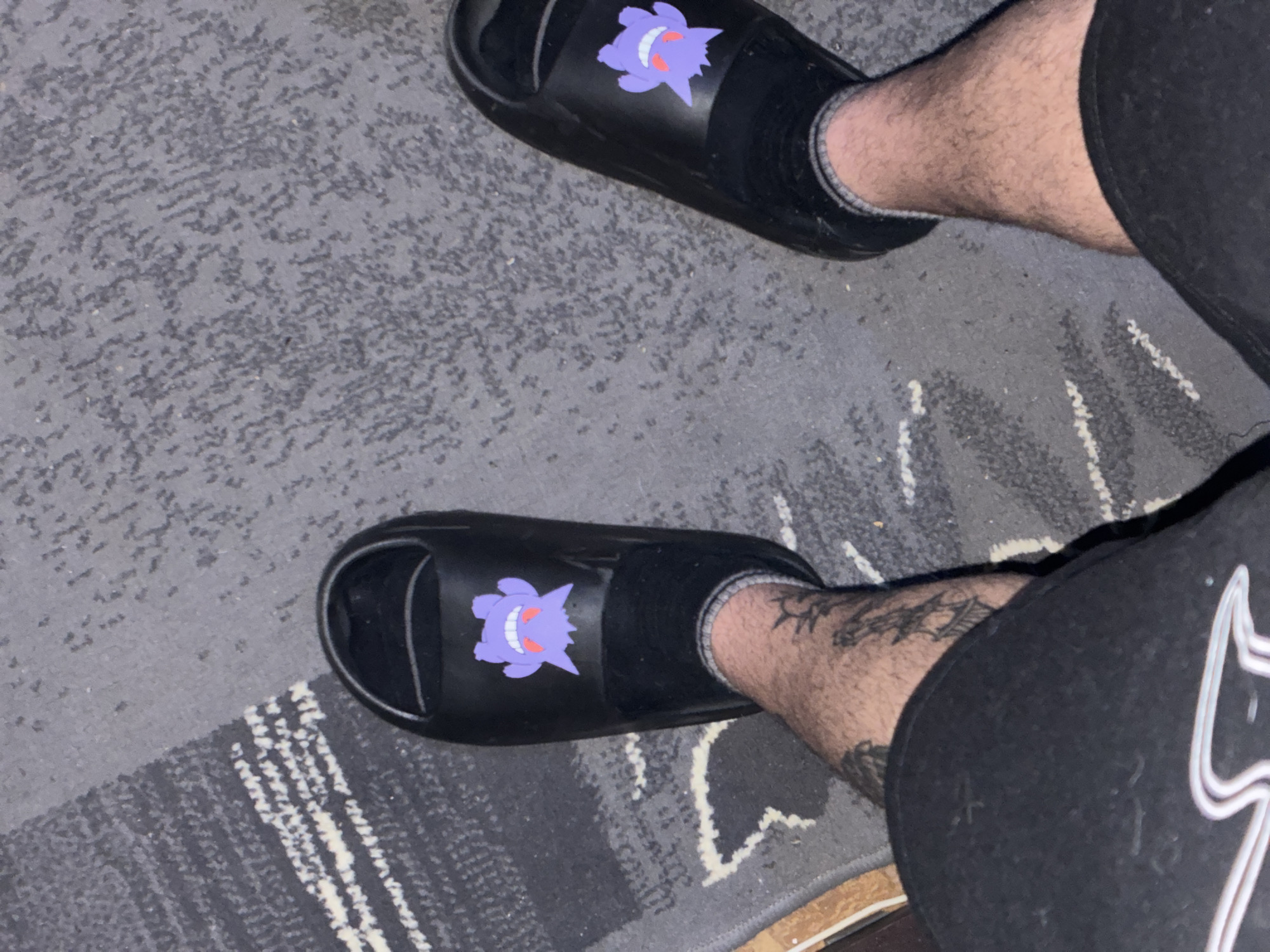 A high-angle shot captures a person's legs and feet wearing black socks and black slides. The slides have a purple Gengar Pokémon character on top. The person is wearing black shorts. The scene is set indoors on a grey and white patterned carpet. The location is unknown and not verifiable as Elhovo, Bulgaria.