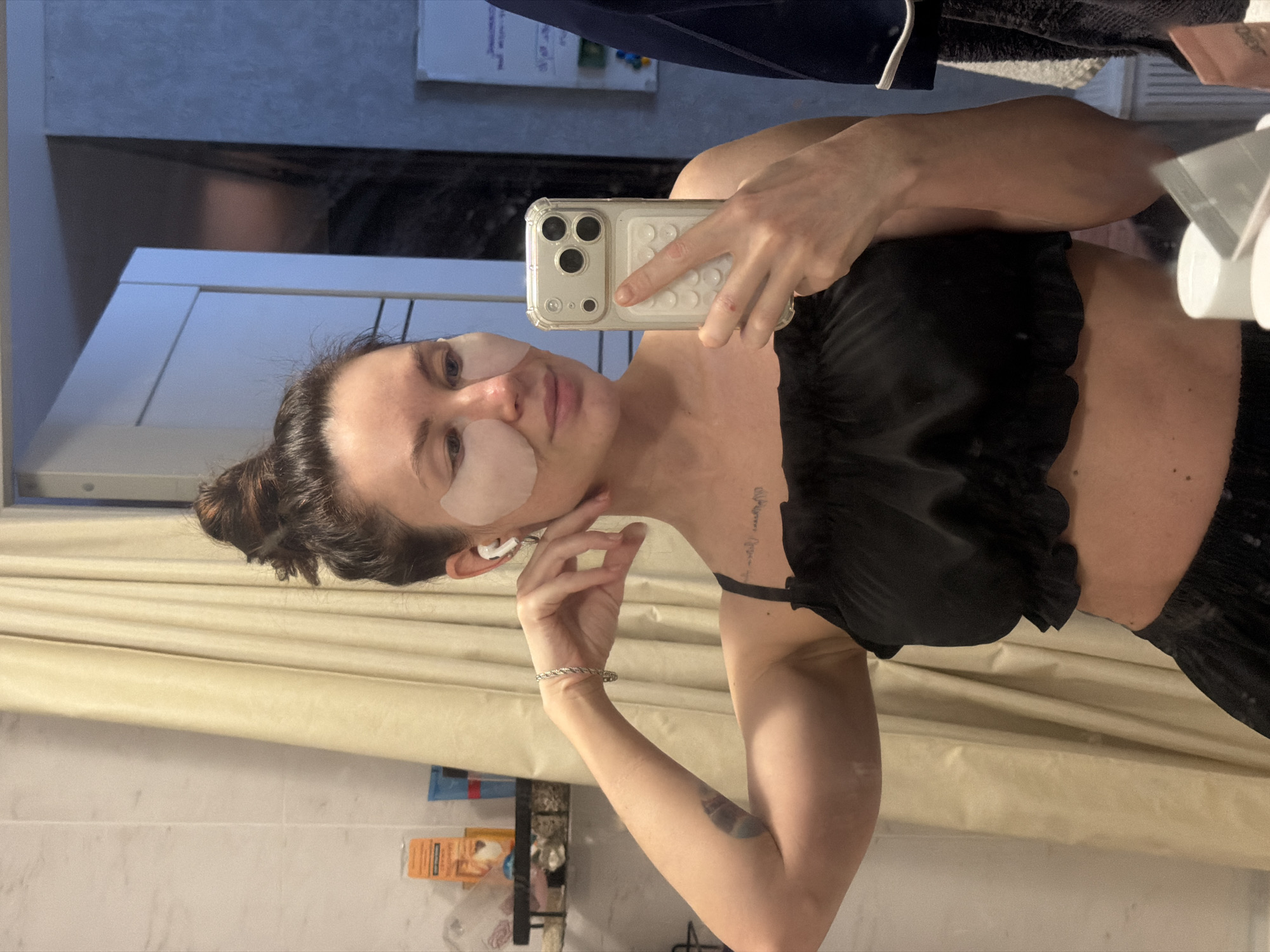 This mirror selfie, taken in a setting that appears to be a bathroom, likely in Perm, Russia, is rotated 90 degrees clockwise. The main subject is a woman with dark hair tied back, who is applying under-eye patches. She is wearing a black ruffled crop top and a white earbud in her left ear. Her right hand holds a light-colored smartphone with a clear, textured case, pointed at the mirror to capture her reflection. A silver-toned twisted bracelet is on her left wrist, and a small tattoo is visible on her left forearm, with a longer, cursive tattoo on her upper left chest/shoulder.
The background shows a beige curtain partially covering a white tiled wall. To the lower left, a shelf holds various toiletries, including an orange-colored container and a blue tube. The reflection in the mirror also shows parts of a dark garment or towel in the upper right corner. The lighting is artificial and bright, indicating an indoor setting. The woman maintains a neutral, relaxed expression during her self-care activity.