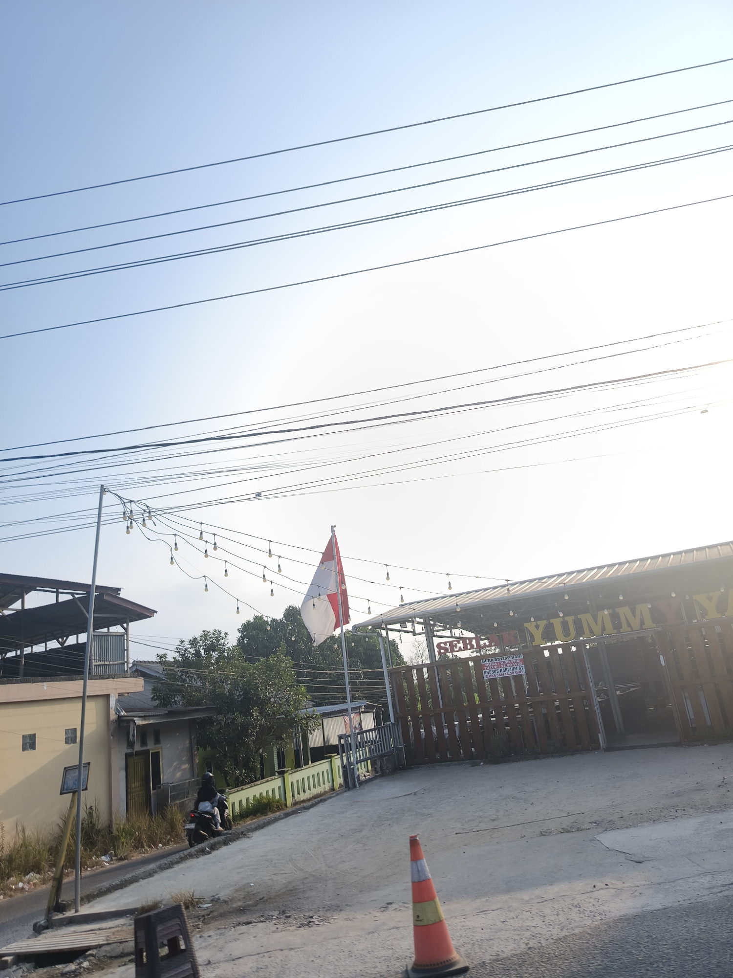 The image depicts a street scene in Pangkalpinang, Indonesia, under a bright, clear sky with multiple power lines stretching across the frame. In the foreground, a road surface is visible, with a single orange traffic cone positioned on the right side. To the left, a man is riding a motorcycle on a sloped road. Behind the road, a building with a yellow facade and a corrugated metal roof is situated. Further back, there are trees and a fence. In the middle ground, a wooden-fenced structure with a roof is present, and an Indonesian flag is flying from a pole to its right. A string of lights hangs from a pole in front of the wooden structure. A sign with text, partially obscured, is attached to the fence. The overall impression is of a semi-urban environment on a sunny day.