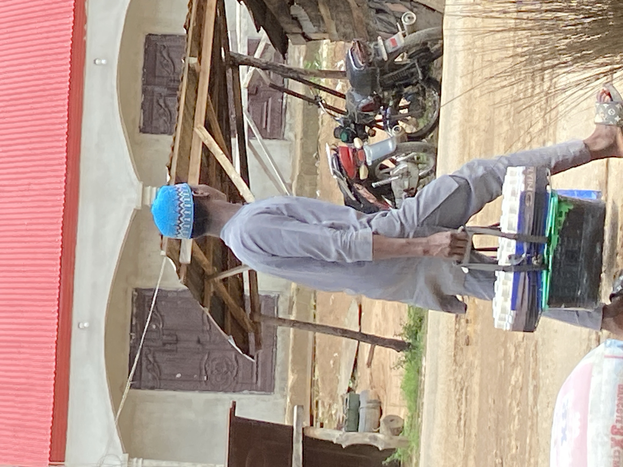 A man wearing a light blue skullcap, a gray shirt, and gray pants is seen carrying plastic containers in Mararaba, Nigeria. He is wearing sandals on his feet. A motorcycle is parked nearby. The backdrop includes a building with wooden framed windows and what appears to be a partially constructed wooden structure adjacent to the building. A bag is partially visible in the foreground.