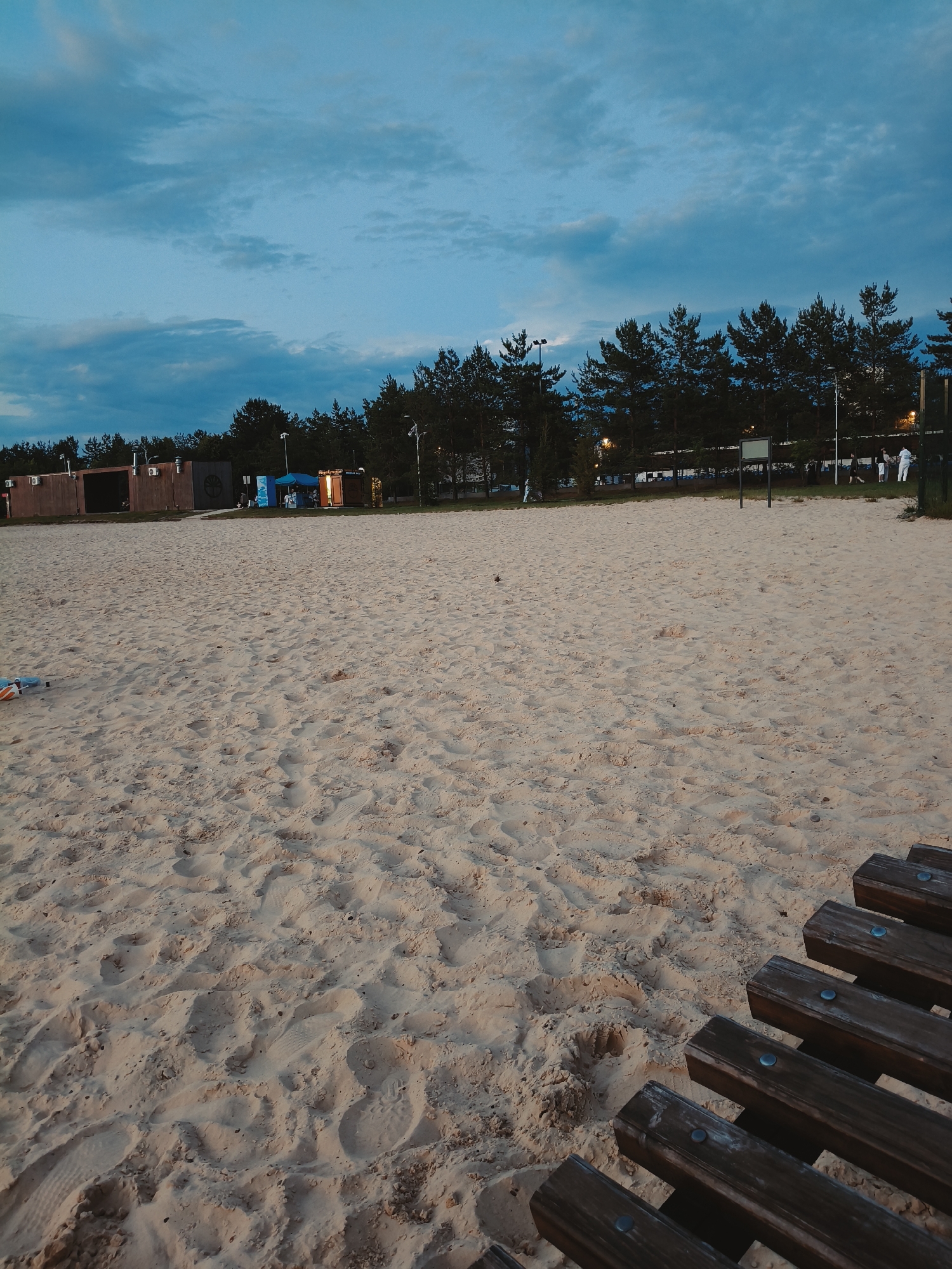 The image depicts a sandy beach area with a line of pine trees in the background, under a twilight sky with scattered clouds. Several wooden structures, possibly small shops or changing rooms, are visible along the edge of the beach. A few people are present near these structures. In the foreground, a wooden bench is partially visible, facing away from the viewer. The sand shows footprints, suggesting recent activity. The overall scene is one of a public beach as evening approaches.