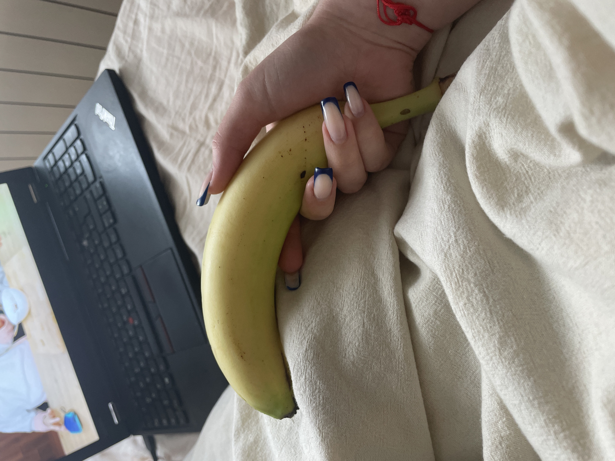 A person is holding a banana in their left hand, with the fingers curled around the fruit, near a black laptop. The person's nails are manicured with a nude base and a dark blue tip. A red string bracelet is visible on their wrist. The background consists of a light-colored, textured fabric. The laptop screen displays an image of a person wearing a white lab coat and mask. The setting appears to be indoors, possibly on a bed or couch. There is no indication of the city of Zlatograd, Bulgaria.