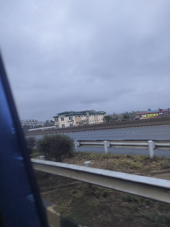 A view from a moving vehicle shows a highway with guardrails. Across the highway is a large building with a light-colored facade and green roof, likely an office complex. To the left, a few apartment buildings are visible. A small bush is positioned between the guardrails and the highway. The sky is overcast. The foreground shows the interior of the vehicle, with a blue window frame visible.