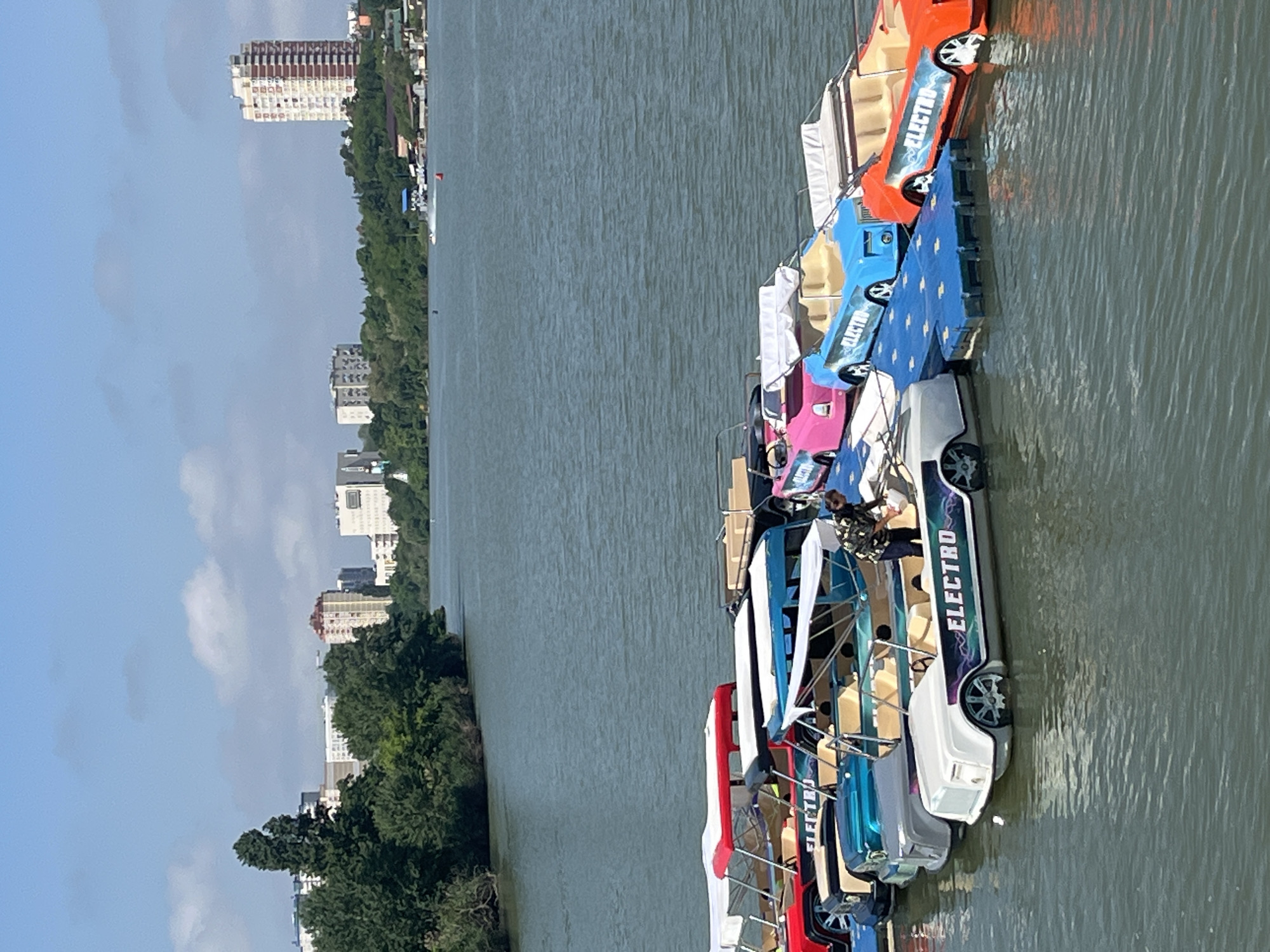 A horizontal medium shot shows a line of docked electric boats on a river, with a forested shoreline and a city skyline in the background. The boats, facing right, are brightly colored (red, blue, orange, pink, white) and have the word "ELECTRO" prominently displayed on their sides. One boat is occupied by a person wearing a harness. The river occupies the majority of the frame, with subtle ripples on the water's surface. On the left side of the frame, a tree-lined bank stretches along the river, with buildings visible in the distance against a slightly cloudy sky. The location is the city of Krasnodar, Russia.