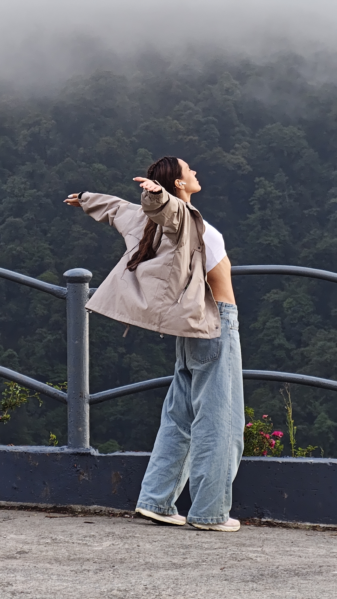 A woman stands on a gray bridge railing overlooking a densely forested mountain range covered in mist, possibly in Binh Lu, Vietnam. She faces slightly to the right of the frame, with her head tilted back and arms outstretched as if embracing the view. She wears a white crop top, loose-fitting light blue jeans, and a beige jacket that is open and flowing behind her. Her dark hair is braided back from her face. The bridge railing is dark gray and made of metal. The background is filled with trees and fog.