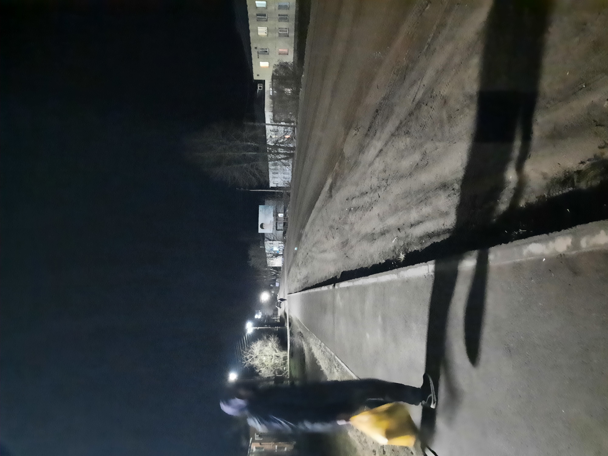 A street scene at night in Krasnogorskoye, Russia, shows a dimly lit street with a sidewalk. Streetlights illuminate the path. A pedestrian is walking on the sidewalk, carrying a yellow bag. A tall building can be seen in the background. The street appears wet, and there is a shadow cast on the sidewalk.