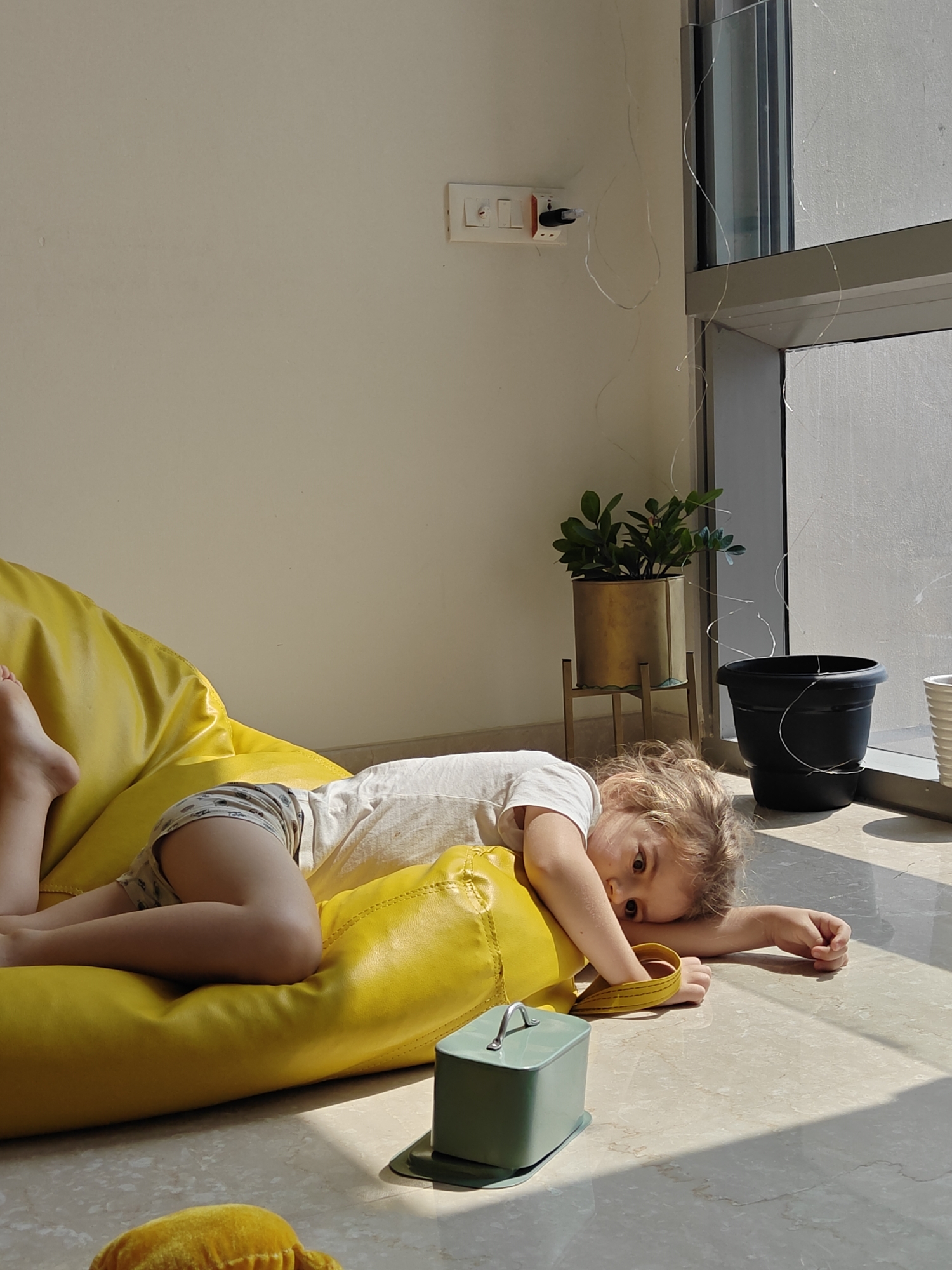 A young child is lying on a yellow beanbag chair indoors in Mumbai, India. The child wears a white t-shirt and patterned shorts and rests their head on their arm. A small, pale green box with a handle sits near the beanbag. Sunlight streams in through a nearby window, illuminating the floor. Two potted plants stand near the window. An electrical outlet is visible on the wall. Thin, decorative lights are draped around the window frame and plants. The floor appears to be marble or tile.