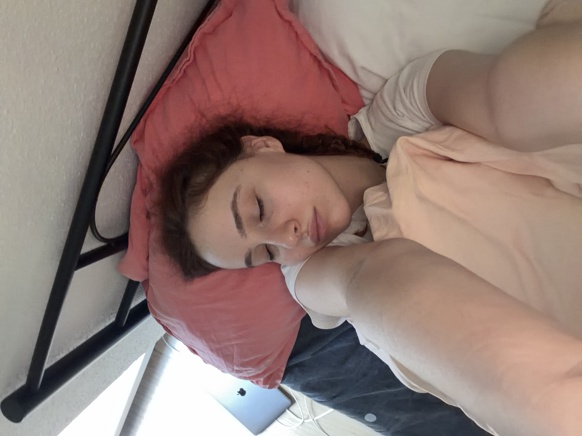 A close-up, high-angle photograph depicts a young adult female resting in a bed in Strasbourg, France. The subject, with fair skin and reddish-brown hair styled in braids, is lying on her left side, facing slightly towards the camera. Her eyes are closed, and her lips are slightly parted. She is wearing a white short-sleeved top.
Her head rests on a salmon-pink rectangular pillow. A white pillow is partially visible to her right. She is covered by a light peach-colored blanket or sheet over her upper body and arms, and a dark blue/grey blanket or duvet over her lower body. A black, tubular metal bed frame is visible along the top and upper left edge of the image, positioned against a textured, off-white wall.
To the left of the bed, on a light-colored surface resembling a wooden floor, a closed silver laptop is present. White cables are visible near the laptop on the floor. The scene is brightly illuminated, with a strong light source, likely a window, visible in the upper left background. The overall environment is an indoor setting, consistent with a bedroom, depicted during daylight hours.