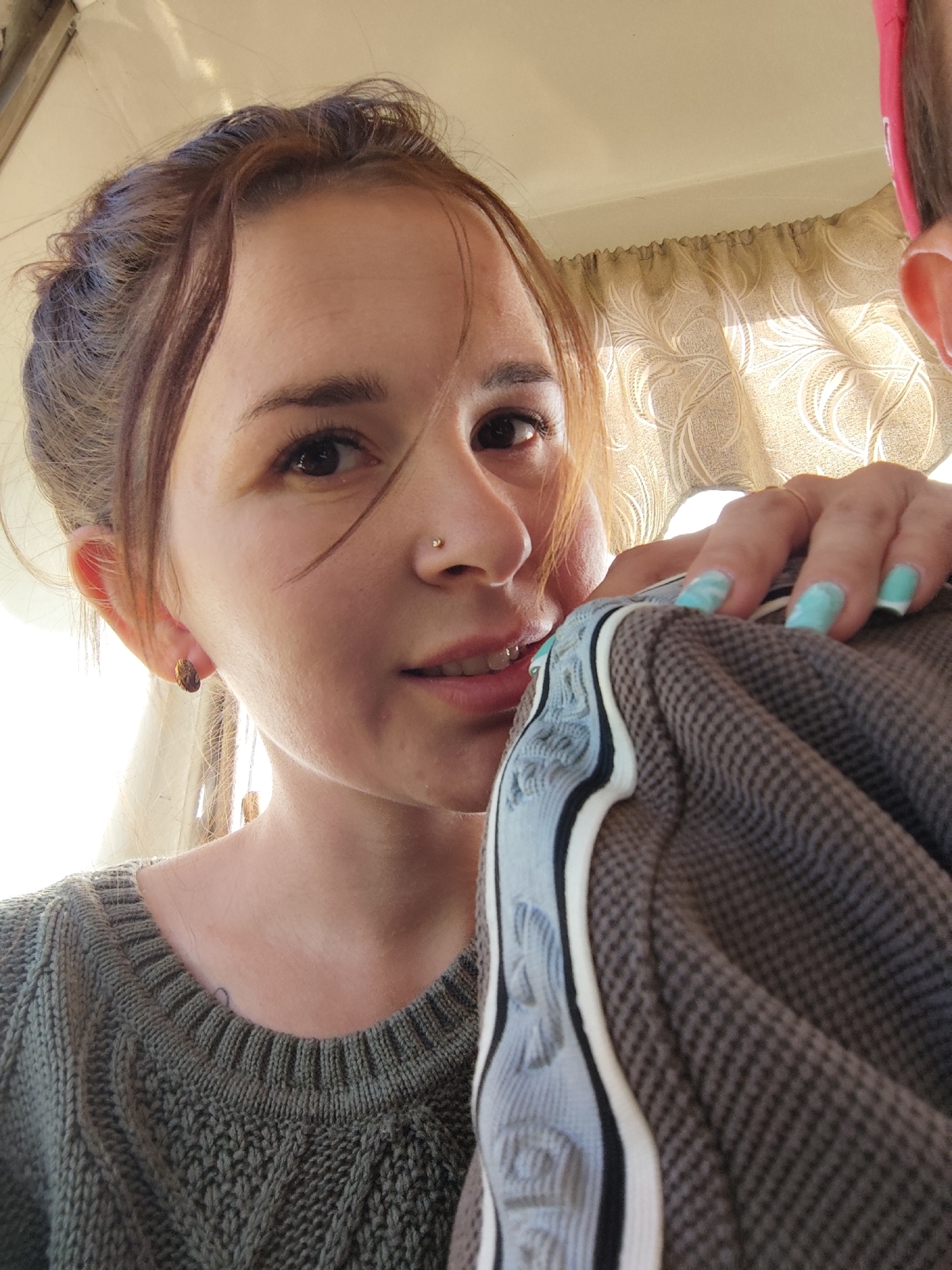 A close-up selfie of a young woman, taken from a slightly low angle. She has light skin, brown hair pulled back with some strands framing her face, and dark eyes. A small stud earring is visible in her left ear, and she has a small nose piercing. She is wearing a dark green knit sweater. She is holding a folded gray and white patterned jacket close to her face, her hand visible with fingernails painted a light blue color. The background shows a patterned curtain and a glimpse of another person with a pink headband, partially out of frame. Based on the request, this scene might be in the city of Chaikovsky, Russia.