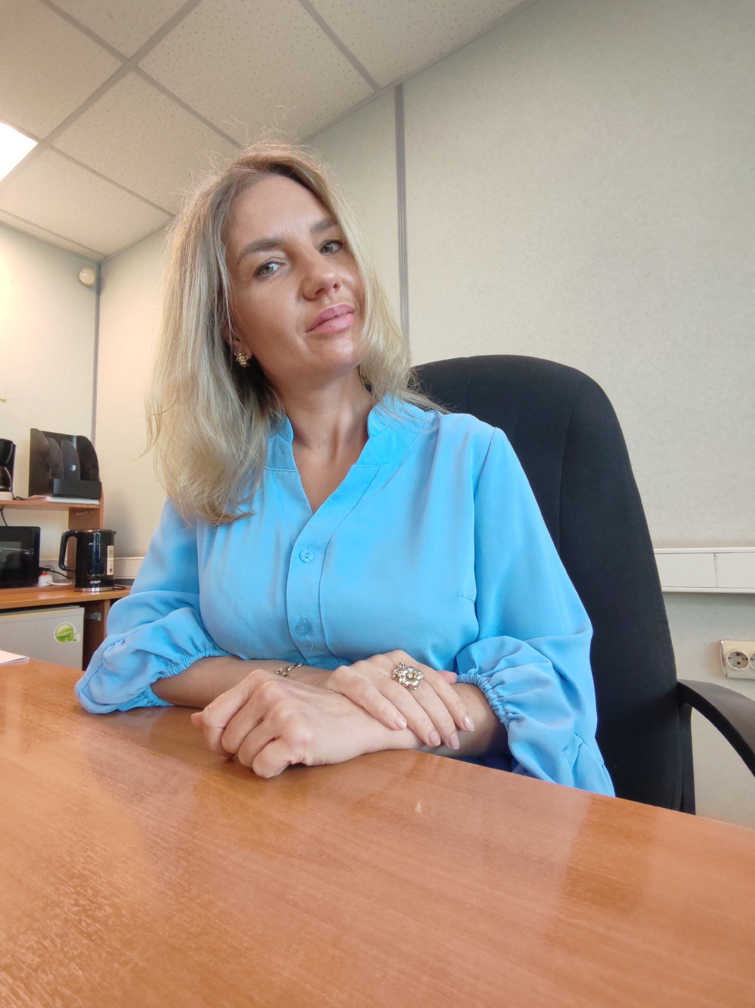 A blonde woman with fair skin and light makeup is seated at a wooden desk in an office. She is wearing a light blue collared blouse with the sleeves rolled up and gathered at the elbow. Her hands are clasped on the desk, and she is wearing a large, decorative ring on her left hand. The background shows a section of an office environment with a black office chair, a light-colored wall, and a ceiling with acoustic tiles and fluorescent lighting. There are also some office supplies visible on a shelf in the background, including black document holders, a black electric kettle, and a small white refrigerator. The overall scene suggests a professional office setting.