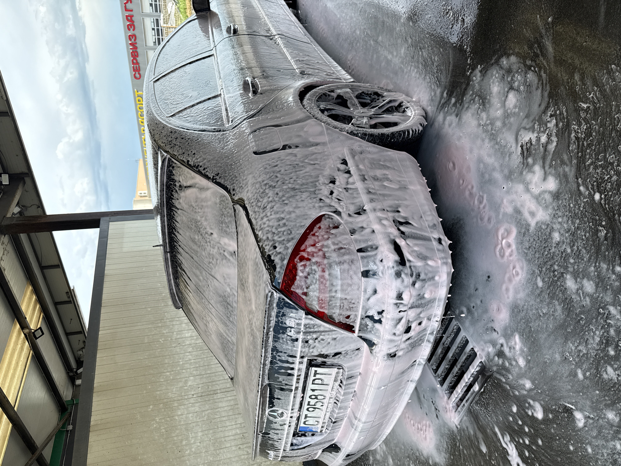 A dark-colored sedan, likely a Mercedes-Benz, is depicted mid-wash at a car wash, heavily covered in white and slightly pinkish cleaning foam. The image is rotated 90 degrees clockwise, making the car appear to be on its side. The vehicle's rear driver's side is prominently visible, showcasing a taillight, a multi-spoke alloy wheel, and a Bulgarian license plate. The license plate reads "CT 9581 PT", along with the country code "B" and EU stars. Below the main number, smaller Cyrillic text states "Всичко само за вашето удобство!" (Everything just for your convenience!).
Foam streams off the car onto the dark, wet ground, accumulating around a metal grate drain visible on the right side of the frame. In the background, beneath a cloudy daytime sky, parts of a building are visible. A large sign on the building displays Cyrillic text: "СЕРВИЗ ЗА ГУМИ И АВТОАКСЕСОАРИ" (SERVICE FOR TIRES AND AUTO ACCESSORIES) and partially "ДИАГНОСТИКА РЕМОНТ" (DIAGNOSTICS REPAIR), indicating an auto service center. The immediate setting includes metal beams and light-colored corrugated panels, typical of a car wash bay. The "CT" on the license plate confirms the location as Stara Zagora, Bulgaria. The scene captures the dynamic process of a car being cleaned at an auto service facility.