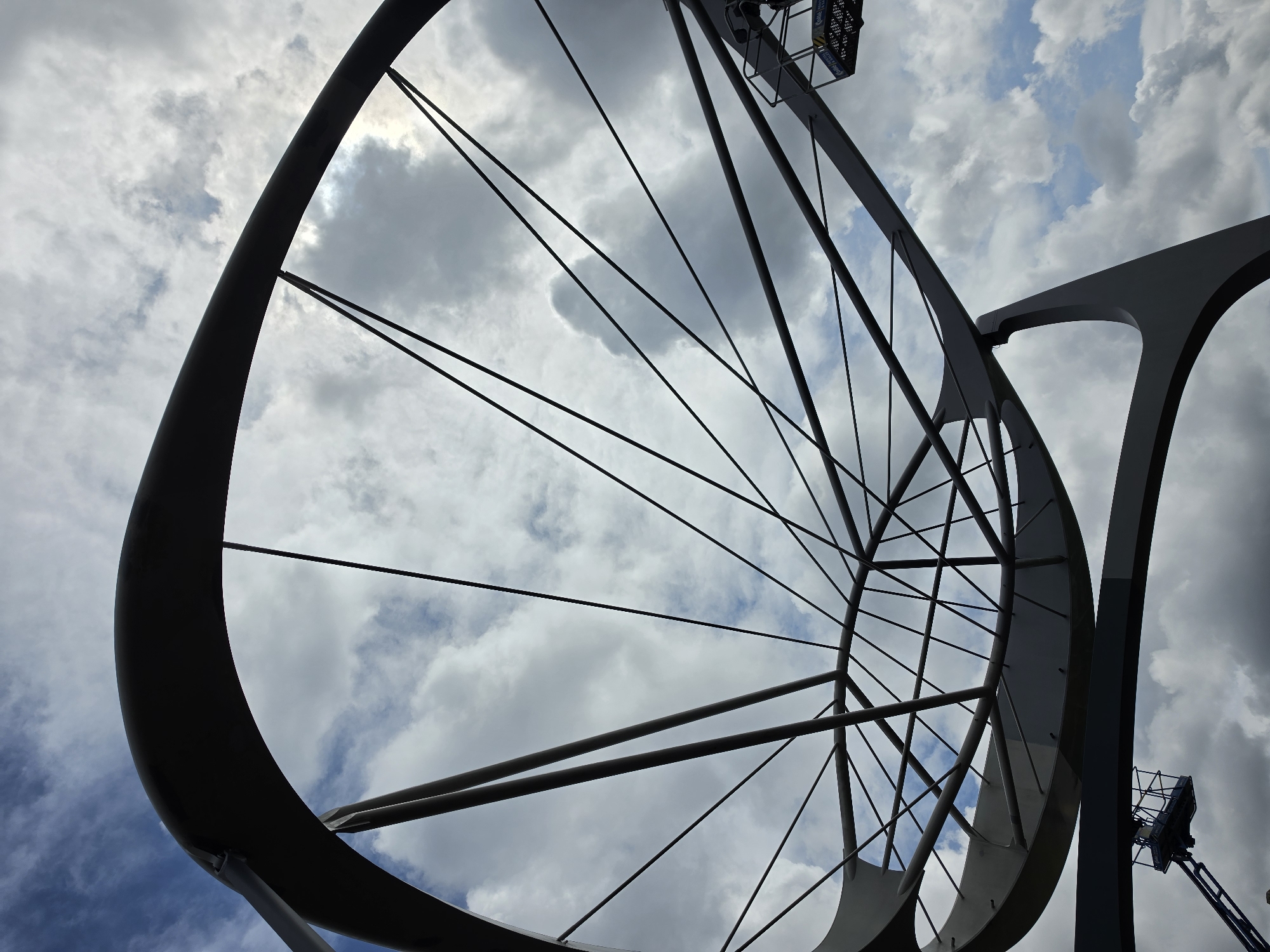 The image shows a low-angle shot of the OVO sculpture in Delft, Netherlands, against a cloudy sky. The sculpture is a large, abstract metallic structure that resembles a stylized wheel or a bird's wing. Thin metal struts radiate from a central point, forming a spoked pattern within a large, curved outer frame. The sculpture is gray and appears to be made of steel. In the lower left and right corners of the image, parts of a blue scissor lift are visible, suggesting that maintenance or construction work may be taking place. The sky is filled with a mix of white and gray clouds, with patches of blue sky visible. The overall impression is of a modern, geometric artwork set against a dynamic natural backdrop.