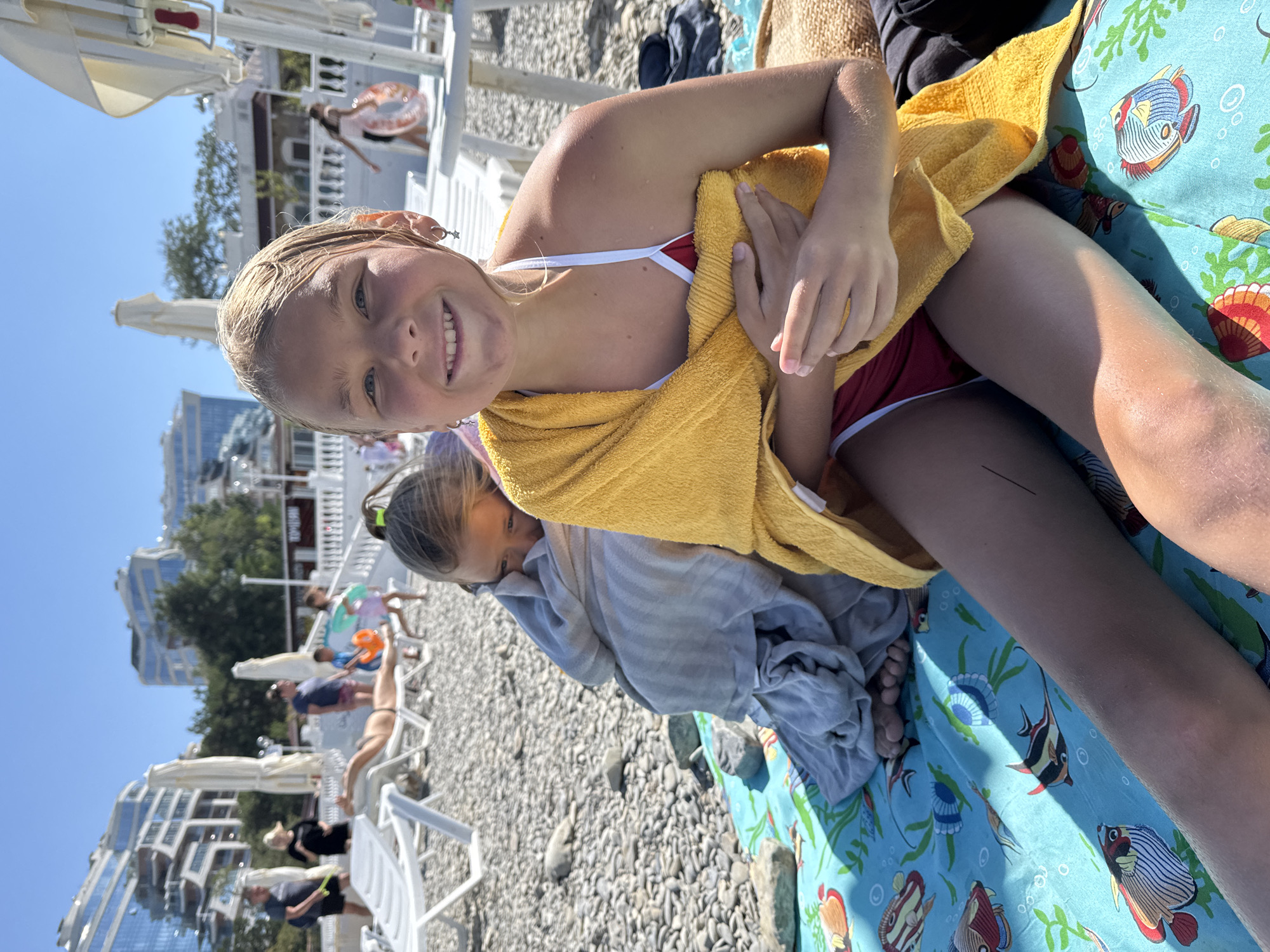 This is a close-up, vertically oriented, outdoor photograph of two young girls on a beach. The girl in the foreground is a blonde child wearing a red and white bikini, wrapped in a yellow towel. She is smiling directly at the camera and appears to be the primary subject. Partially visible behind her is another child, also wrapped in a gray towel. They are both lying on a beach blanket decorated with colorful fish and coral.
The setting is a pebbly beach with numerous white lounge chairs and umbrellas visible in the background. Beyond the beach, there are buildings with modern architecture and trees. The sky is clear and blue, indicating a sunny day. The overall atmosphere is relaxed and summery, suggesting a vacation or a day spent at the beach. The time of day appears to be midday due to the bright sunlight and clear skies. There is no visible text in the image. The location context is Gelendzhik, Russia.