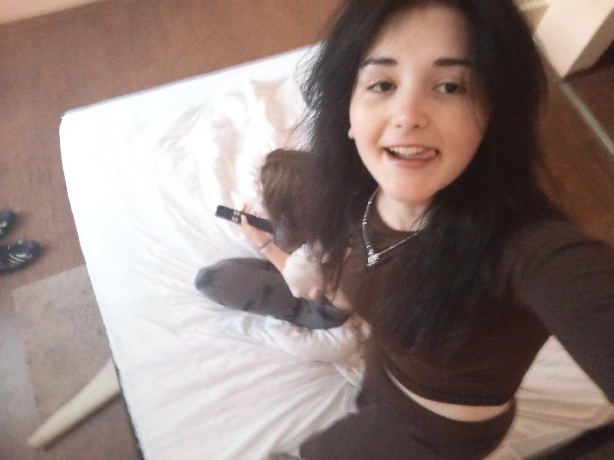 A young woman with dark hair is taking a selfie in a room with a bed. The bed has white sheets, and a second person is partially visible in the background, holding a remote control. The room is indoors, and the location is presumed to be Poltava, Ukraine. Other objects visible include shoes and a portion of a wooden structure.