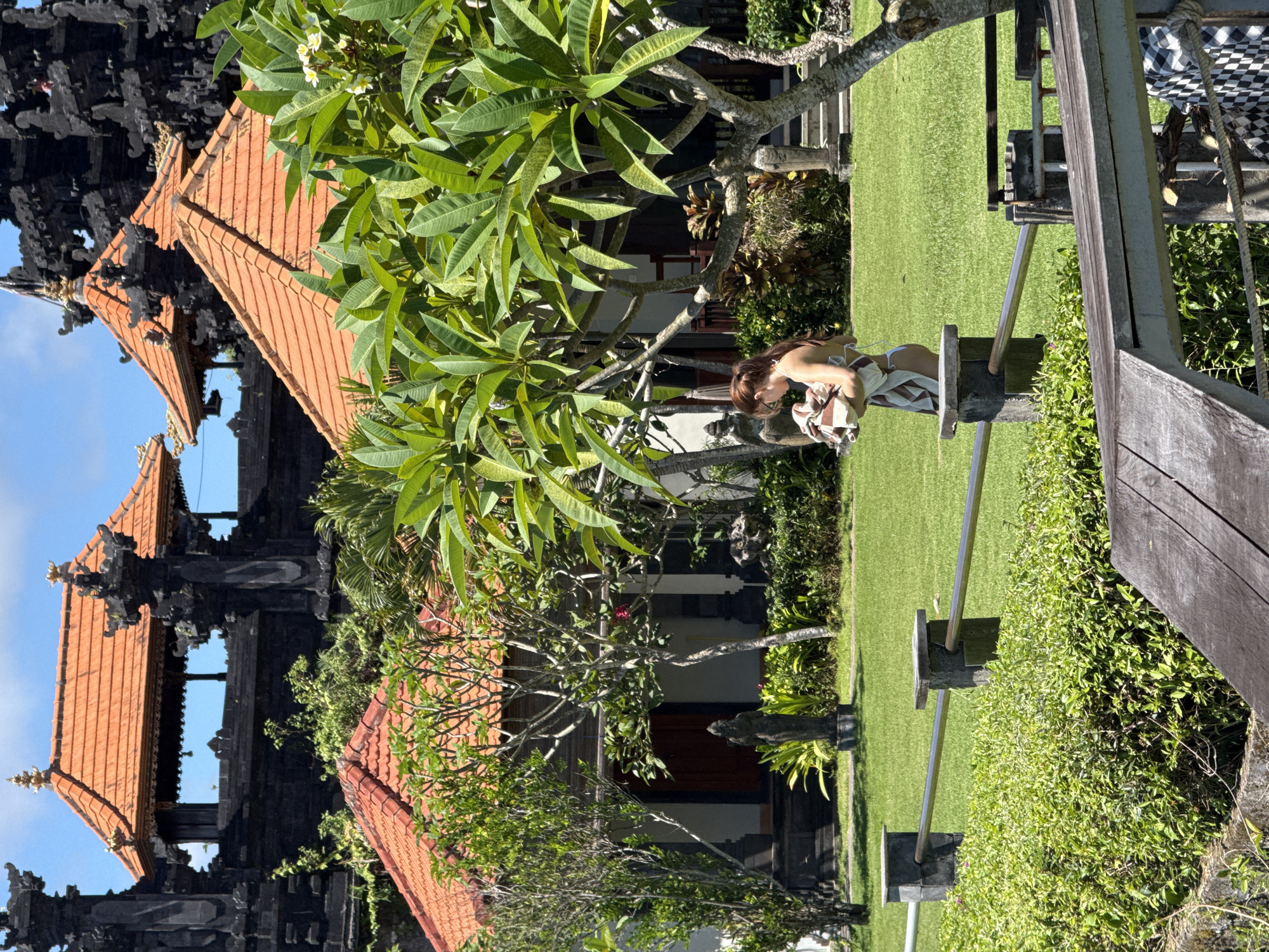 This image captures a serene outdoor scene in Kuta Selatan, Indonesia, bathed in bright daylight under a clear blue sky. The setting features traditional Balinese architecture and lush tropical greenery.
In the mid-ground, a woman with brown hair, wearing a light-colored top and striped bottoms, is standing or sitting near a wooden and metal structure, possibly a swing or bench. She is looking down at an object she holds with both hands.
The background is dominated by distinct Balinese buildings with traditional terracotta-tiled roofs. Some structures are dark and intricately carved, possibly indicating a temple gate or ornate cultural architecture. Various trees with vibrant green leaves, including some with characteristic white frangipani flowers, are interspersed throughout the landscape, adding to the tropical ambiance.
The foreground and mid-ground consist of a meticulously maintained green grass lawn, bordered by dense green shrubs and hedges. Wooden planks and metal supports are visible on the right side of the frame, forming part of the structure near the woman. The overall impression is one of tranquility and cultural richness. No visible text is present in the image.