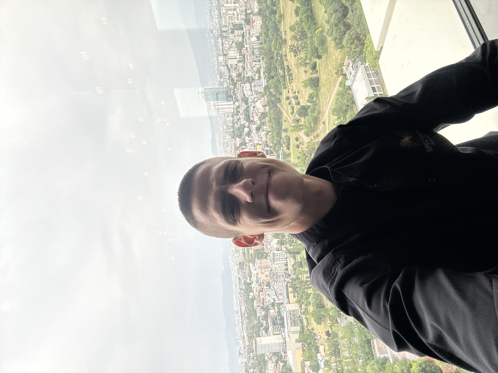 The image is a portrait of a young man in what appears to be a black shirt, taken from a high vantage point, likely a skyscraper in Sofia, Bulgaria. The man is positioned in the foreground, facing the camera and smiling slightly. His short, dark hair is neatly cut.
Behind the man, a sprawling cityscape is visible, indicative of Sofia. Buildings of various sizes and architectural styles are densely packed, interspersed with green spaces that suggest parks or tree-lined streets. The backdrop features a hazy mountain range, a common feature of the Sofia landscape. The sky is overcast, suggesting a cloudy or possibly rainy day.
The man's shirt has a small logo on the left chest area. The visible text on the logo, though partially obscured and blurry, reads "Sberbank". This suggests the man might be an employee or representative of Sberbank. The overall mood of the image is casual and friendly, with the man's relaxed posture and smile contributing to this impression. The time of day is difficult to determine precisely due to the overcast conditions, but it appears to be daytime.