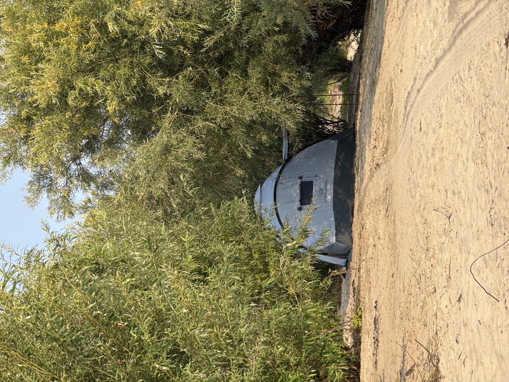 A gray and black tent is set up on a sandy beach, partially obscured by the dense green foliage of trees. The tent has a window and a logo that reads "NORTHLAND". The sandy ground shows tire tracks, indicating recent vehicle activity. The environment appears to be a natural, possibly wooded area near a body of water, consistent with a location in Barsovo, Russia. The sky is clear and blue. There are no people visible in the image.