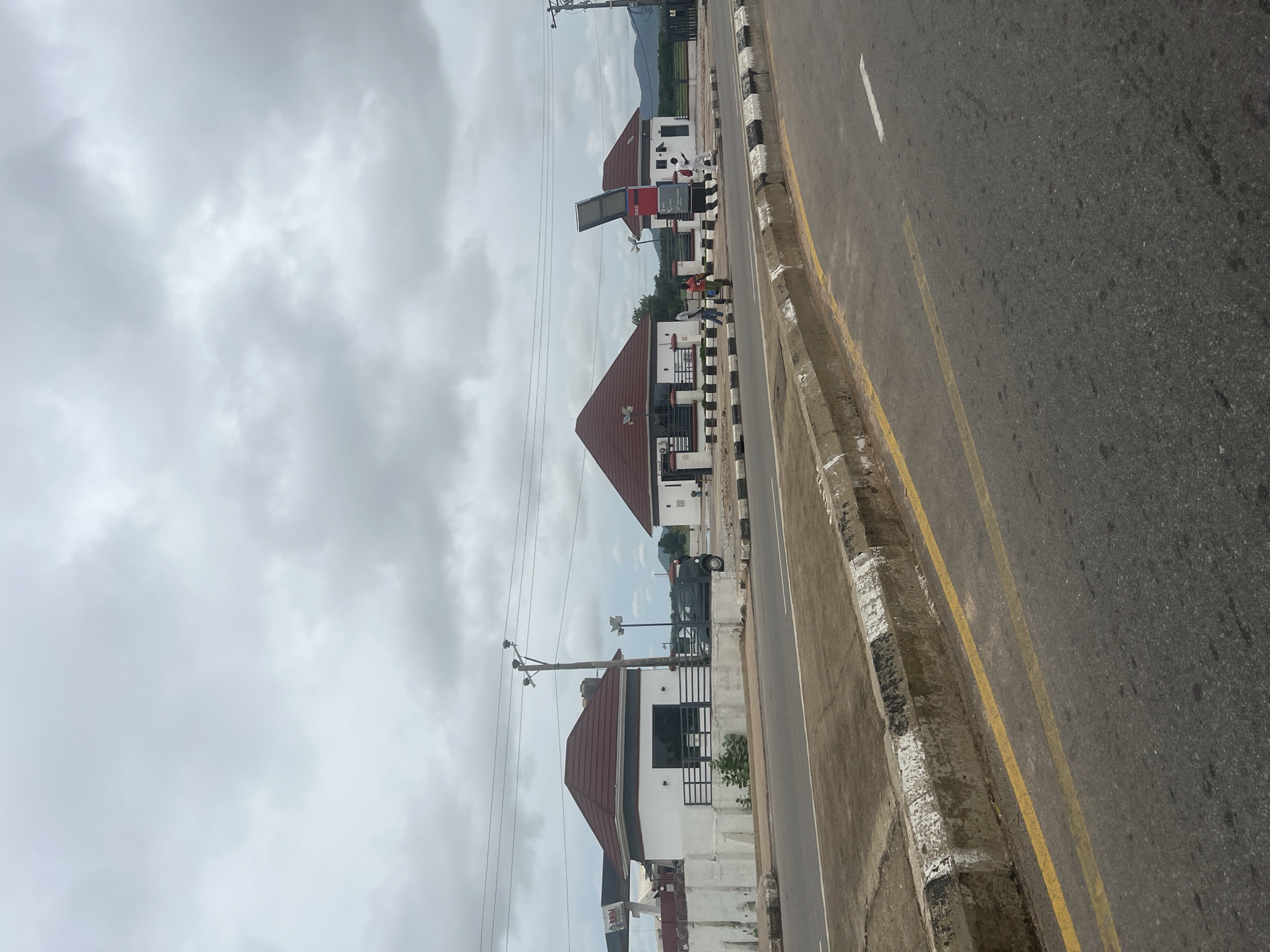 The image depicts a cloudy day in Jalingo, Nigeria, with a focus on a road and several small, modern buildings lining its side. The road surface is asphalt, marked with yellow and white lines, and bordered by a concrete curb with black and white markings.
On the right side of the road, there are two similar, white, one-story buildings with distinctive red, pitched roofs. The building closer to the viewer has a prominent sign that reads "GAS" and appears to be a service station. A solar panel is angled upwards from this structure. To its right, another identical building is visible.
In front of the buildings, there is a paved area with decorative black and white patterned borders. Several people are present: two men in white shirts and red shorts are standing near the gas station, and another man in a blue shirt and dark pants is walking with a handcart. A dark-colored car is parked in front of the leftmost building, which also has a small black metal fence.
The environment appears to be a relatively modern development, with power lines visible overhead, supported by a utility pole. In the background, rolling green hills and more distant mountains are silhouetted against the overcast sky. The overall impression is that of a roadside commercial area under a heavy cloud cover.