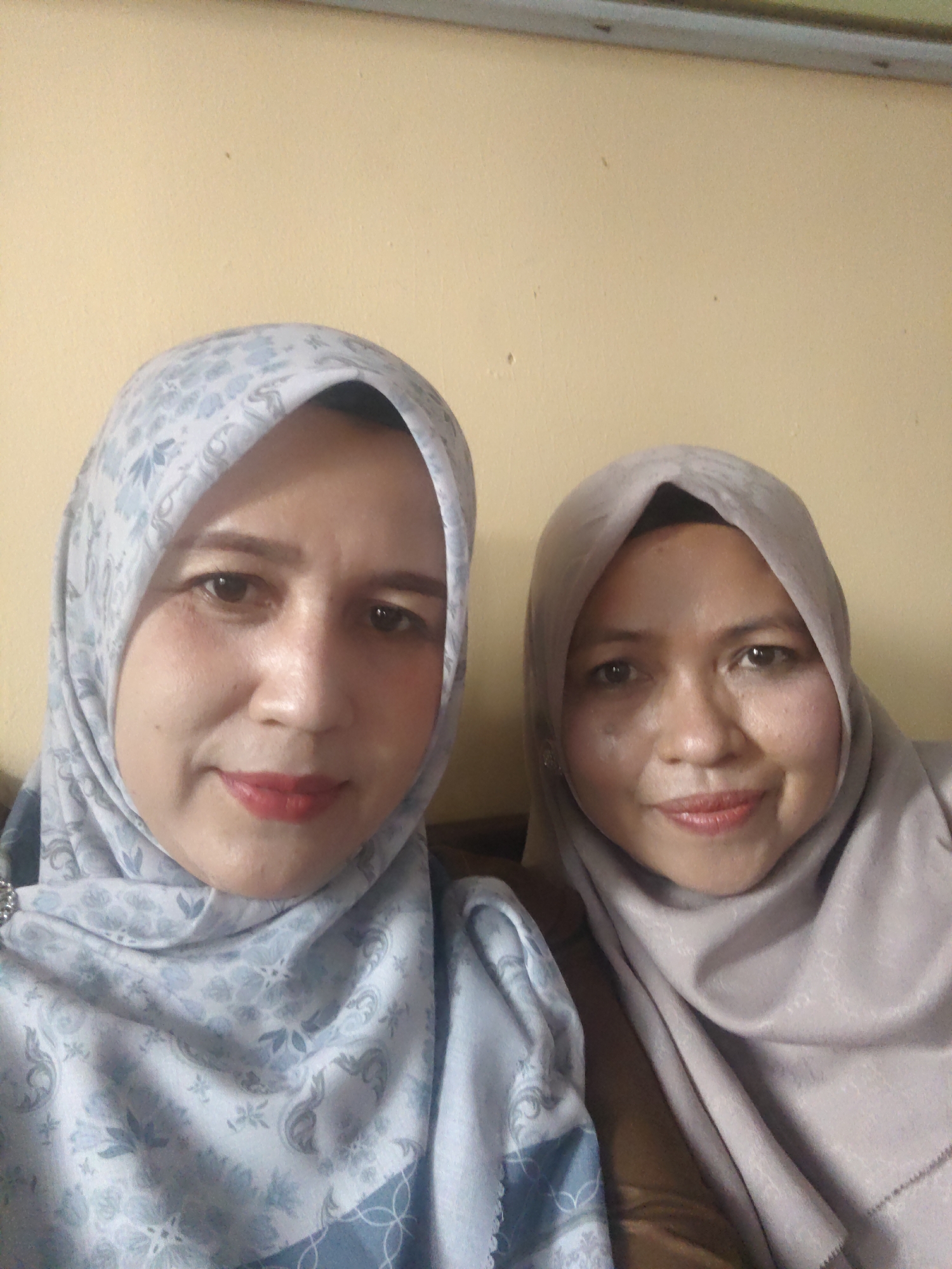 Two women wearing hijabs are posing for a selfie against a light yellow wall. The woman on the left has a light blue hijab with a floral pattern and is wearing light makeup. The woman on the right has a taupe hijab and is also wearing light makeup. The background appears to be a domestic interior. The image was taken in Keudejungkagajah, Indonesia.