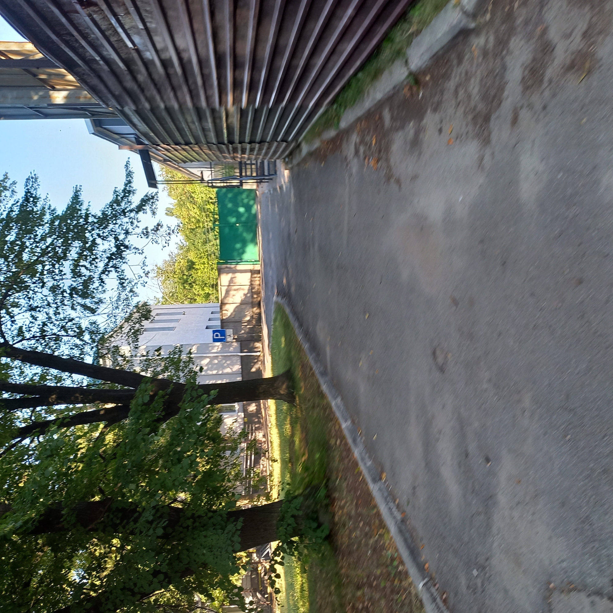 The image depicts a paved pathway with a slight upward incline, leading towards a green gate and a building in the background. To the left of the pathway, there is a grassy area with trees, and a white building with windows. A blue parking sign with a white 'P' is visible on the left side of the pathway, near the building. To the right of the pathway is a dark brown corrugated metal fence. The scene appears to be an outdoor, possibly industrial or administrative, area. The lighting suggests it is daytime, with sunlight casting shadows. No people are visible in the image. The location is identified as Kropyvnytskyi, Ukraine, implying an urban or suburban setting.