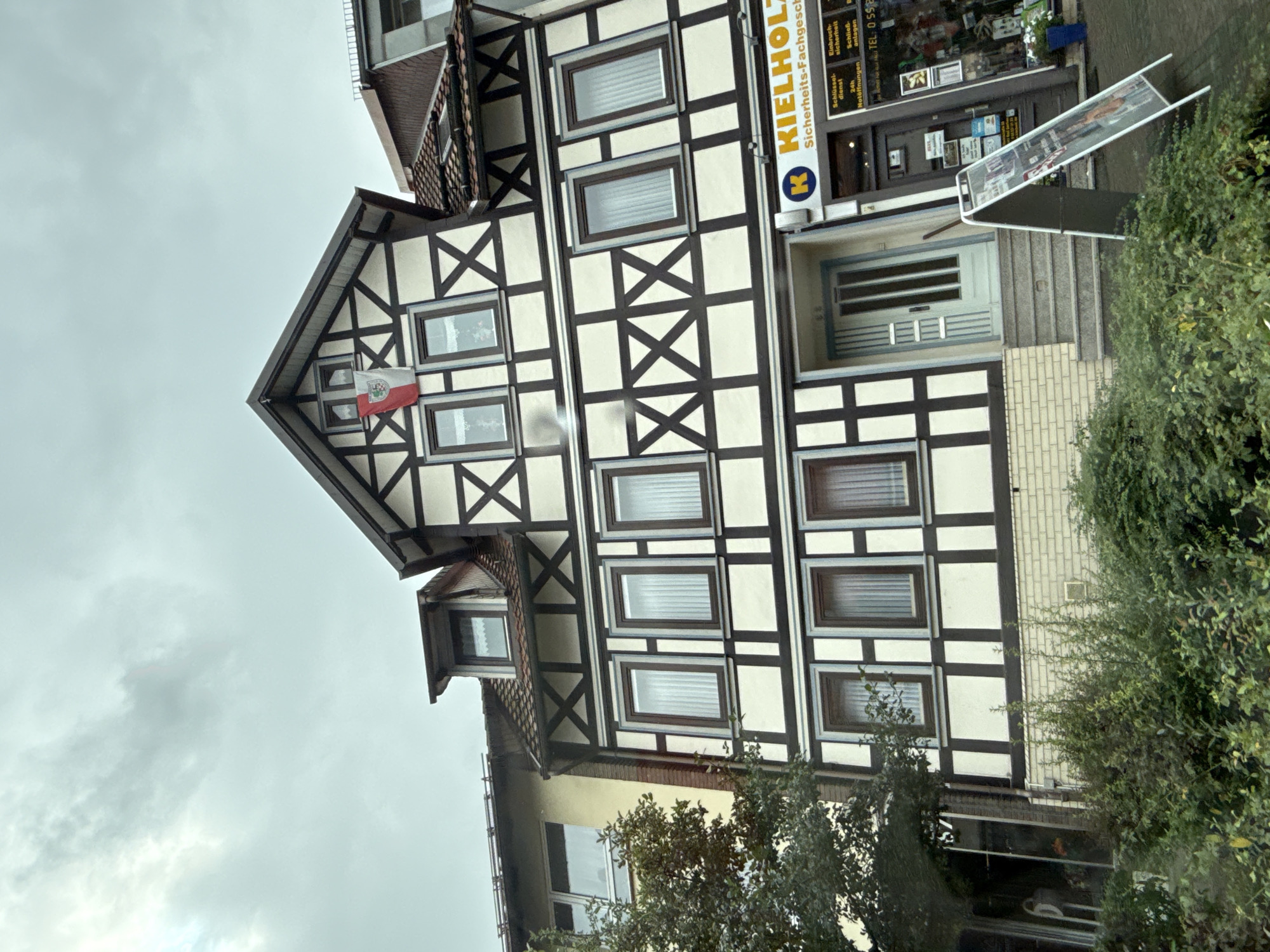 The image shows a three-story half-timbered building in Bad Sachsa, Germany. The building has a traditional facade with white stucco and dark brown wooden beams. A flag with a red and white design and a crest is displayed on the upper story. Below the flag, several windows with white curtains are visible. The ground floor features a light blue door with glass panes and a set of stairs leading up to it. Adjacent to the door is a shop with a blue and yellow sign that reads "KIELHOLZ Sicherheit-Fachgeschaft". An A-frame sign displaying advertisements stands in front of the shop. Green bushes and trees are present in the foreground, and the sky is overcast.