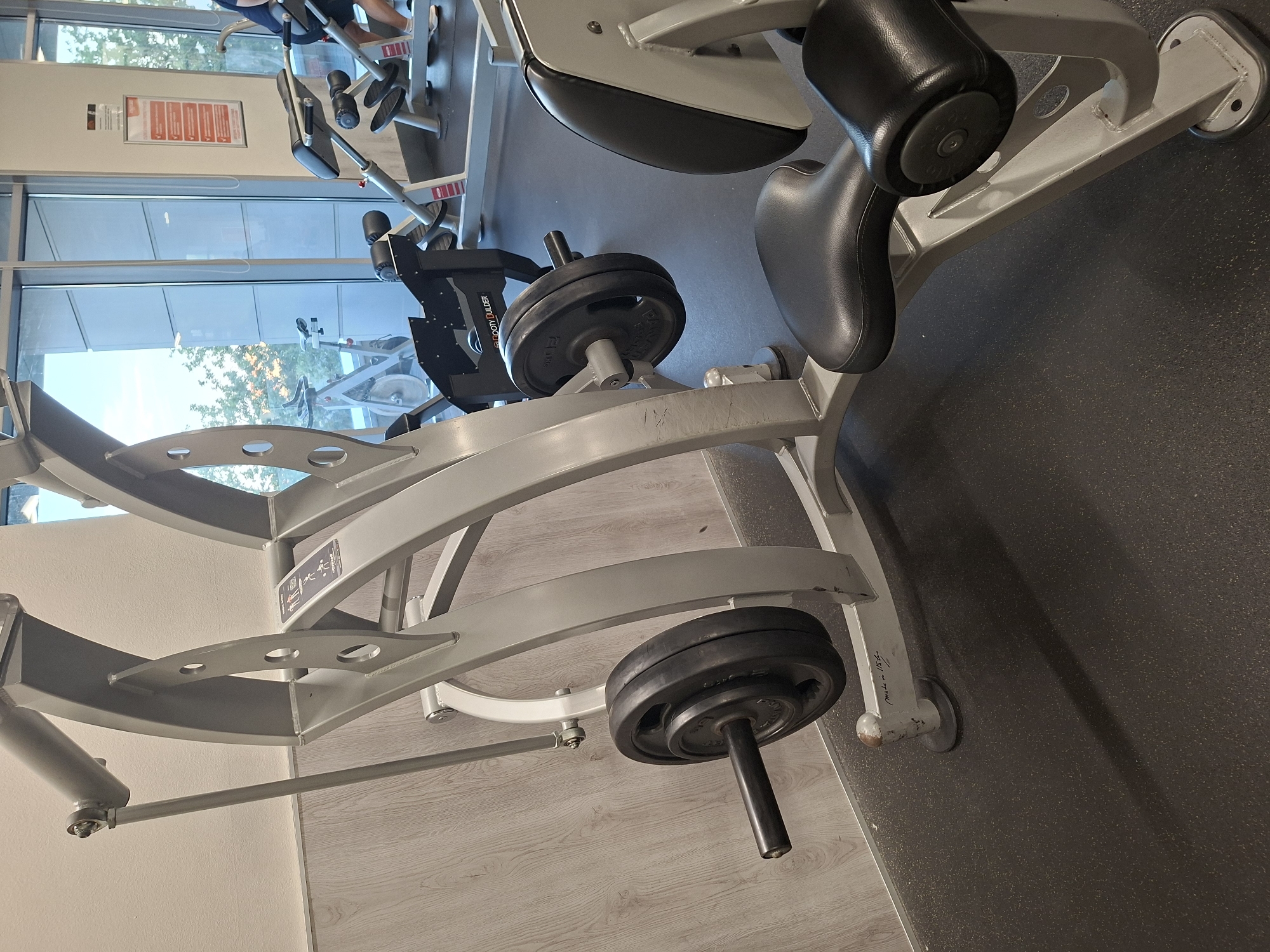 A weight-training machine is shown in a gym. The machine is gray with black padded seats and armrests. Two weight plates are loaded onto a bar attached to the machine. In the background, a person is visible using another piece of gym equipment and a spinning bike is visible through a large window. The floor is black rubber. The walls are white and light wood paneling. The scene is indoors.