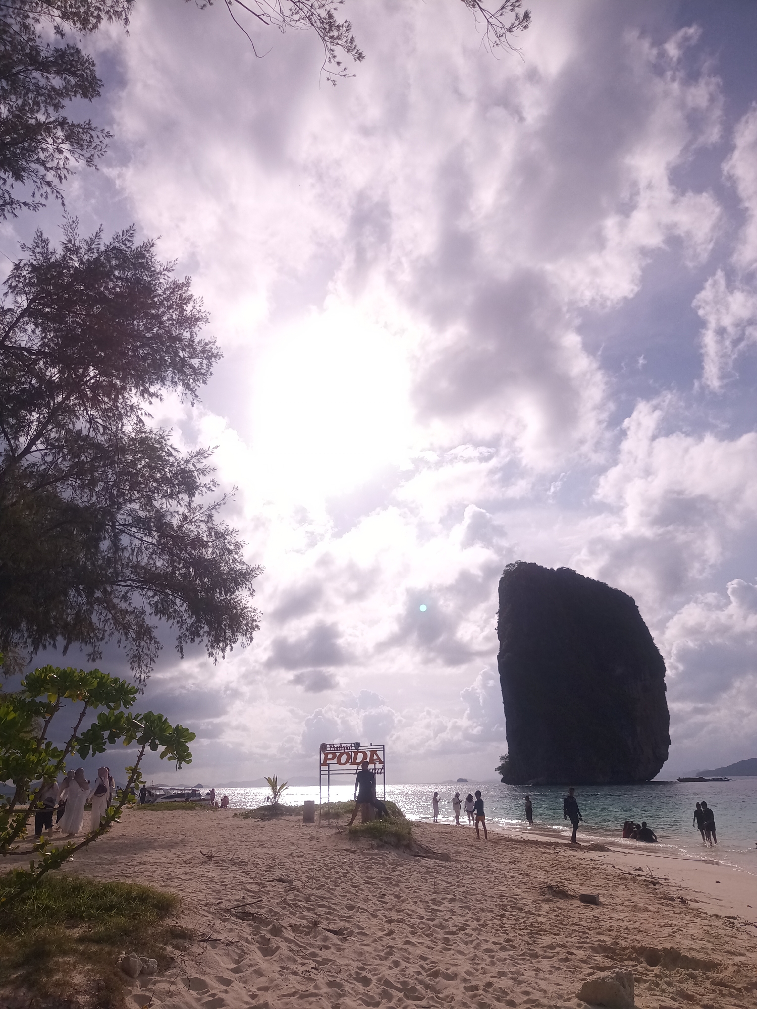 The image shows a beach scene in Ban Ao Nang, Thailand. The sky is filled with clouds and the sun is shining brightly. In the distance, there is a large rock formation in the ocean. On the beach, there are several people, some standing near the water and some are in the water. There is also a sign that says "PODA". There is a tree on the left of the image. There is a boat in the water.