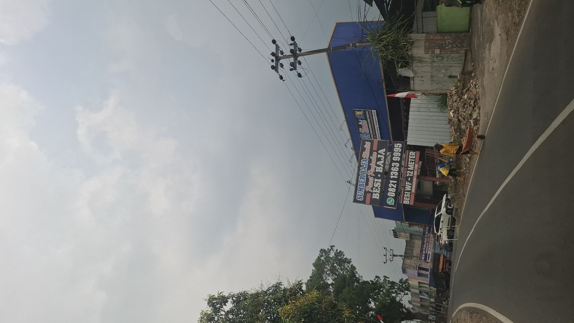 This is a vertical, medium-shot, outdoor photograph taken on a cloudy day in Susukan, Indonesia. The foreground features a grey asphalt road with white lane markings curving to the left. In the mid-ground, a blue building with a white and red sign is visible. The sign advertises "SUMBER BAJA MANDIRI PUSAT PENJUALAN BESI - BAJA" with a phone number "0821 1363 9995" and "Besi WF - 12 Meter". To the right of the building, two men are visible. One man, wearing a yellow shirt and blue pants, is pushing a wheelbarrow filled with debris. Another man, wearing a yellow shirt and yellow boots, is seen behind him, also working with debris. A pile of construction debris, including bricks and rubble, is located on the right side of the road. To the left of the building, a white car and a dark truck are parked. A large tree with green foliage stands on the far left side of the image. Power lines and poles are visible throughout the scene. The background consists of a grey, overcast sky with scattered clouds.