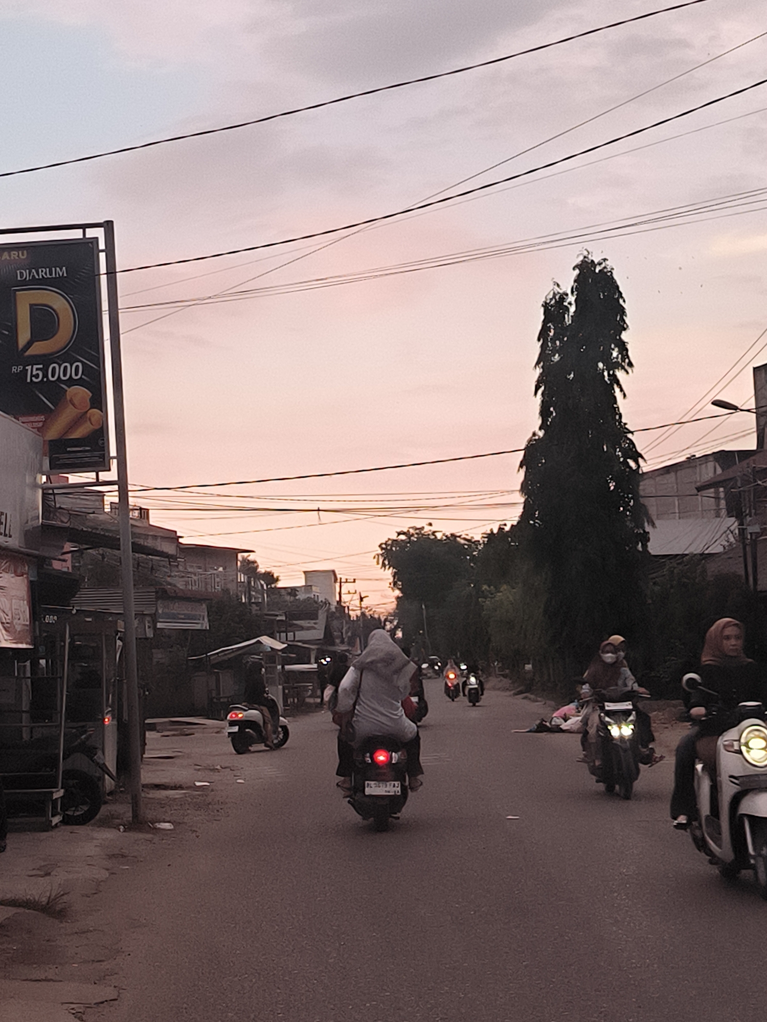The image depicts a street scene in Langsa, Indonesia, at dusk. The sky is a muted pink and gray. Overhead, numerous power lines crisscross the view. In the foreground, several people are riding scooters on the road. One scooter is closest to the viewer, carrying two people, the one on the back wears a head covering. Other scooters are further down the road, also carrying people, some wearing head coverings. A tall, dark green tree stands near the center of the image. Buildings of varying heights and architectural styles line the sides of the street. A large sign with the word "DJARUM" and the price "RP 15.000" is visible on the left side of the frame. The overall impression is a busy urban environment at the end of the day. The road surface appears slightly uneven and dusty.