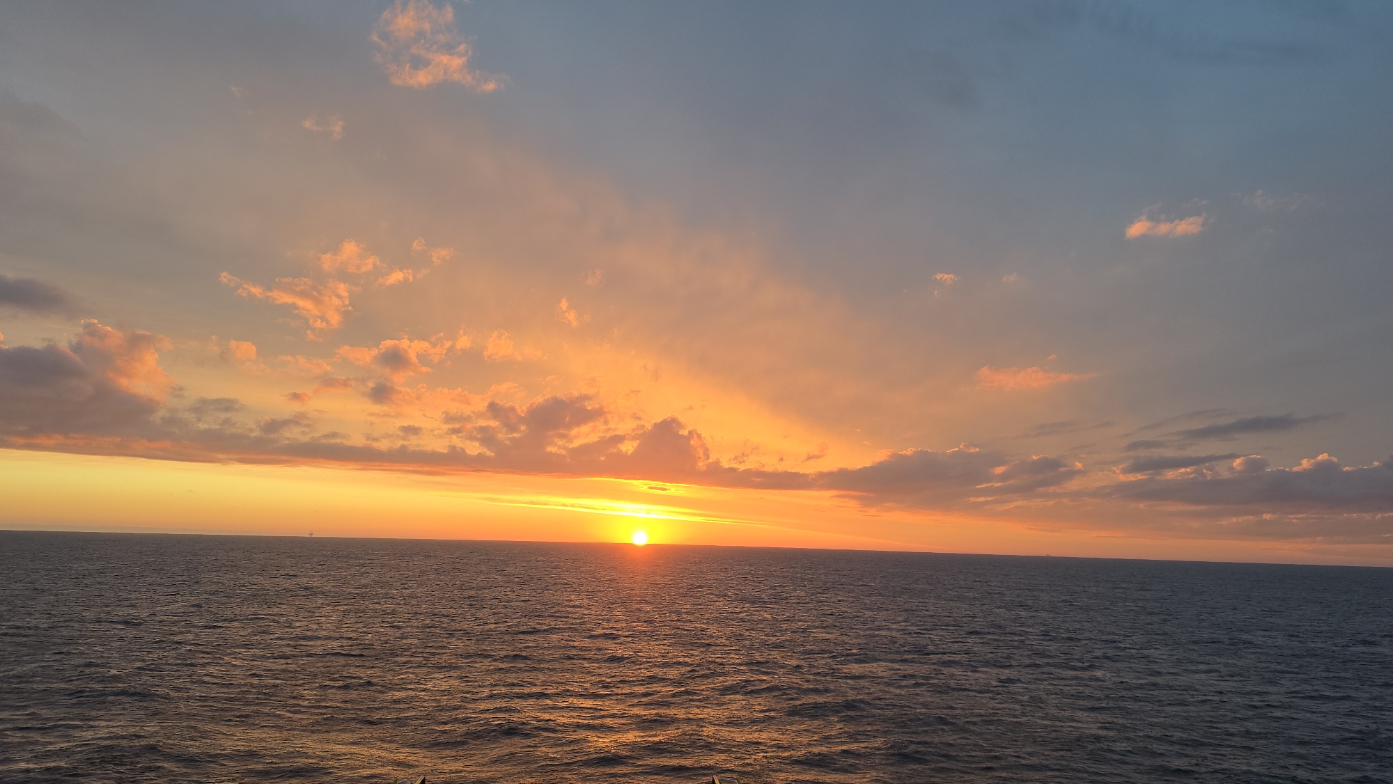 The image shows a sunset over the ocean. The sun is setting on the horizon, with a bright, orange glow. The sky is a mix of orange, yellow, and blue colors. There are some clouds in the sky, which are also reflecting the colors of the sunset. The ocean is dark blue, with some ripples on the surface. The location of this scene is Unknown, Unknown. There are no people or other objects visible in the image. The only action is the sunset. The environment is the ocean.