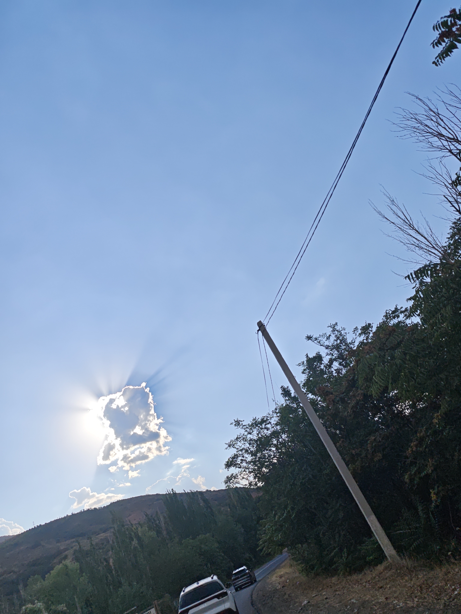 The image depicts a road in Yangikurgan, Uzbekistan, curving through a landscape dominated by hills covered in vegetation. A concrete utility pole stands prominently on the right side of the road, supporting overhead power lines that traverse the frame. The sky is bright blue with a single, small cloud partially obscuring the sun, which creates a crepuscular ray effect. Dense green foliage lines the right side of the road, with trees also dotting the slopes of the hills on the left. Two cars are visible on the road, one in the foreground and another further in the distance, suggesting movement. The overall scene captures a rural or semi-rural setting on a clear day.