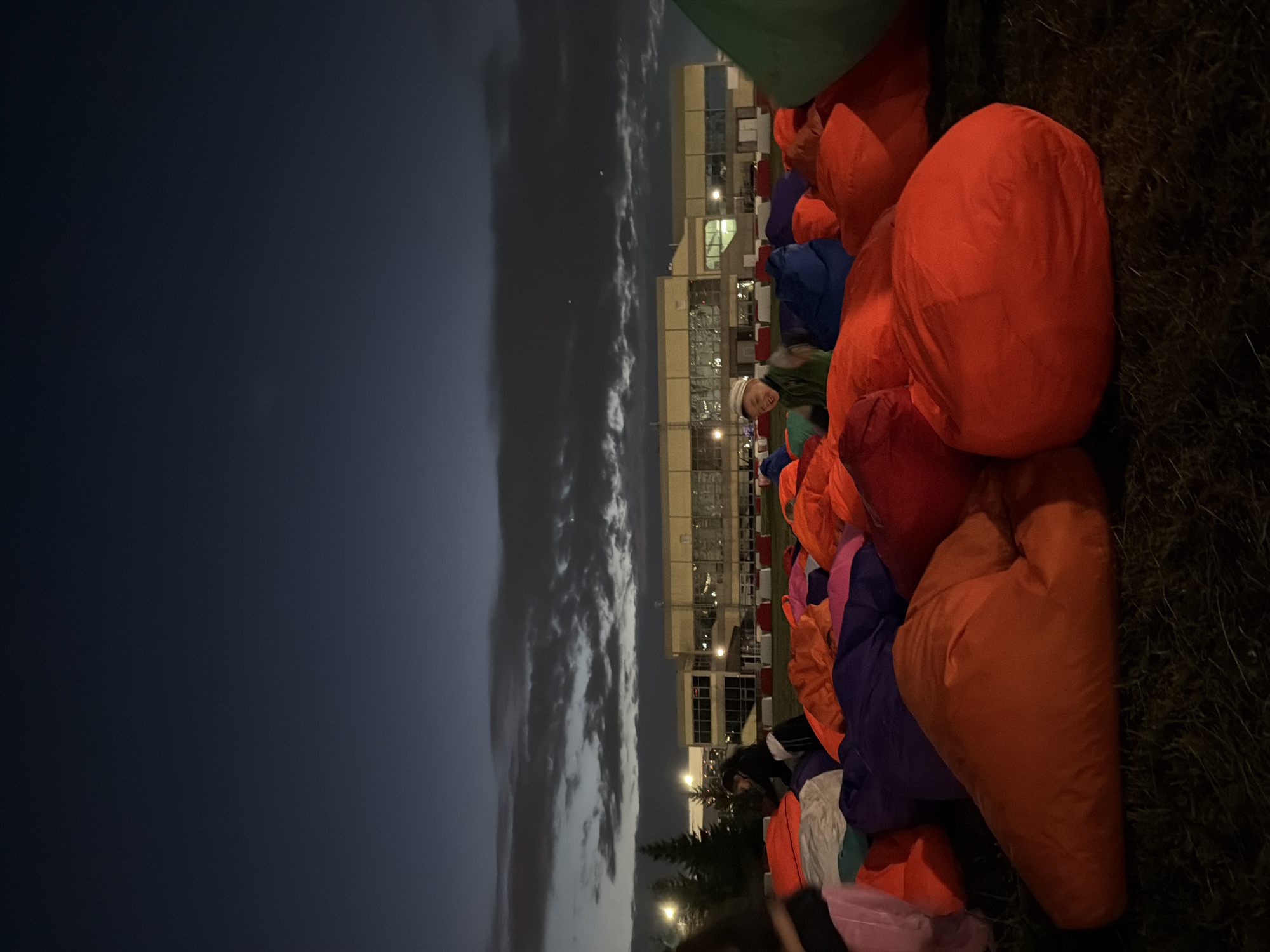 An outdoor scene at dusk depicts a gathering of people sitting on a large field of beanbag chairs. The beanbag chairs are brightly colored in hues of orange, purple, green, and pink, creating a vibrant and playful atmosphere. In the background, a modern building with large windows is visible, possibly a school or community center, with lights illuminating its interior. Trees line the edge of the field, and a cloudy sky with hints of the setting sun adds to the ambiance. One person, a young boy wearing a hat and a vest, is seated on a beanbag chair and looking towards the camera with a smile. Other individuals are partially visible, some interacting with each other, while others are obscured by the beanbag chairs. The overall scene suggests a casual outdoor event, possibly a movie screening or a community gathering, set against a backdrop of a darkening sky and urban architecture.